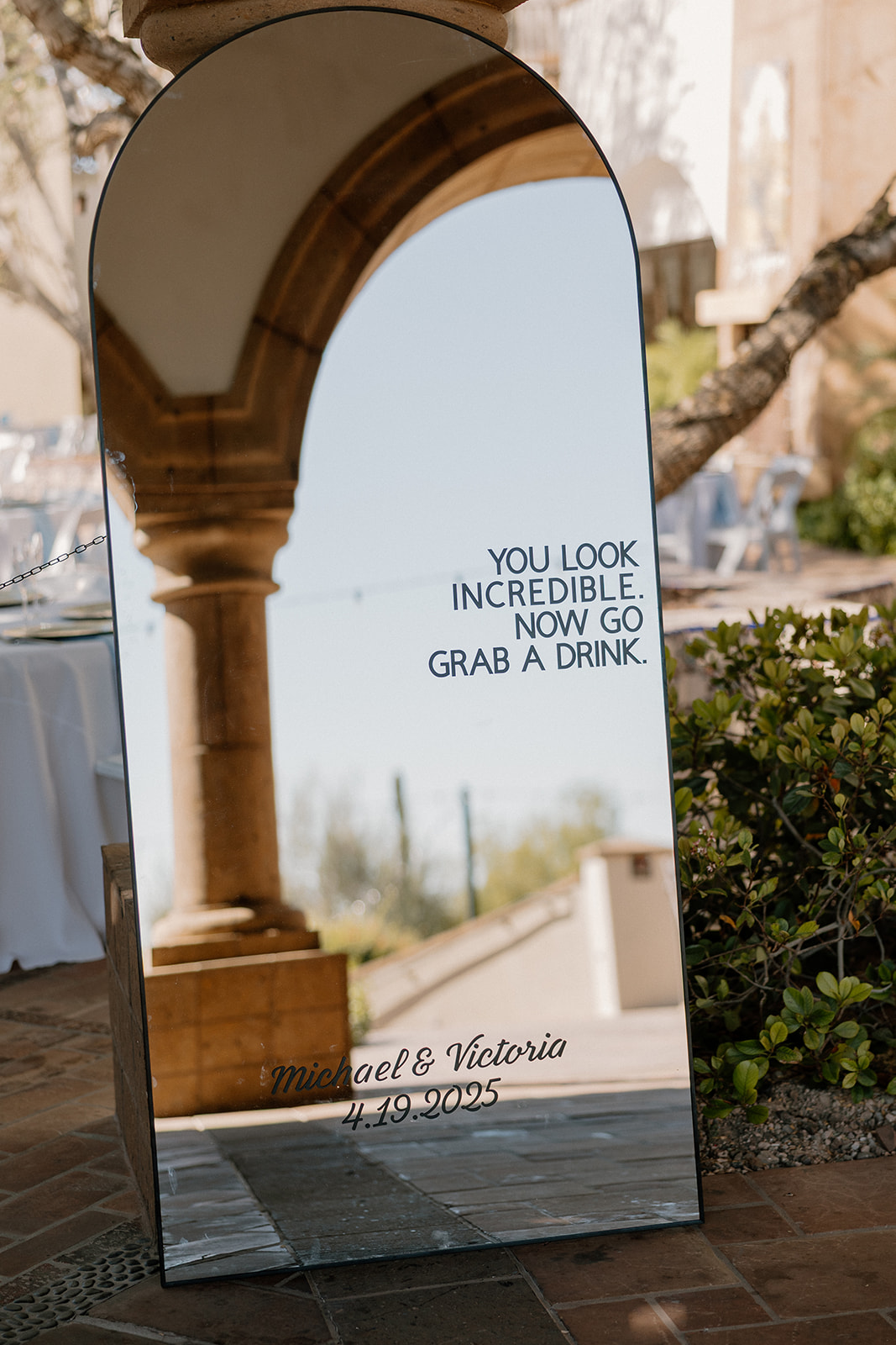 Modern mirror sign at cocktail hour reads “You look incredible. Now go grab a drink.” with the couple’s names and date etched below.