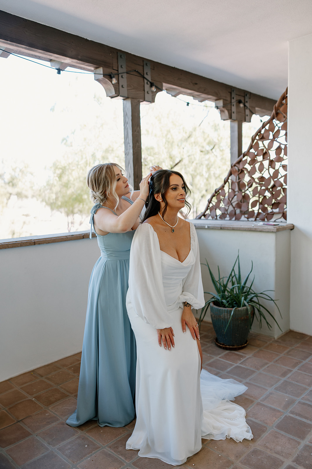 A bridesmaid in dusty blue helps the bride with final touches before the ceremony, sharing a quiet, focused moment.