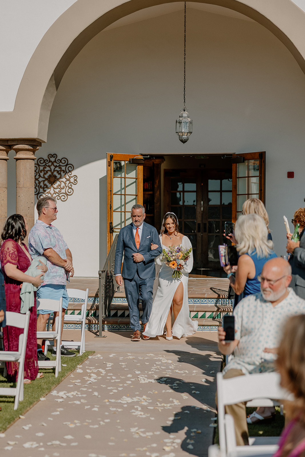 The bride walks down the aisle arm-in-arm with her father, guests rising in anticipation at the start of the ceremony.