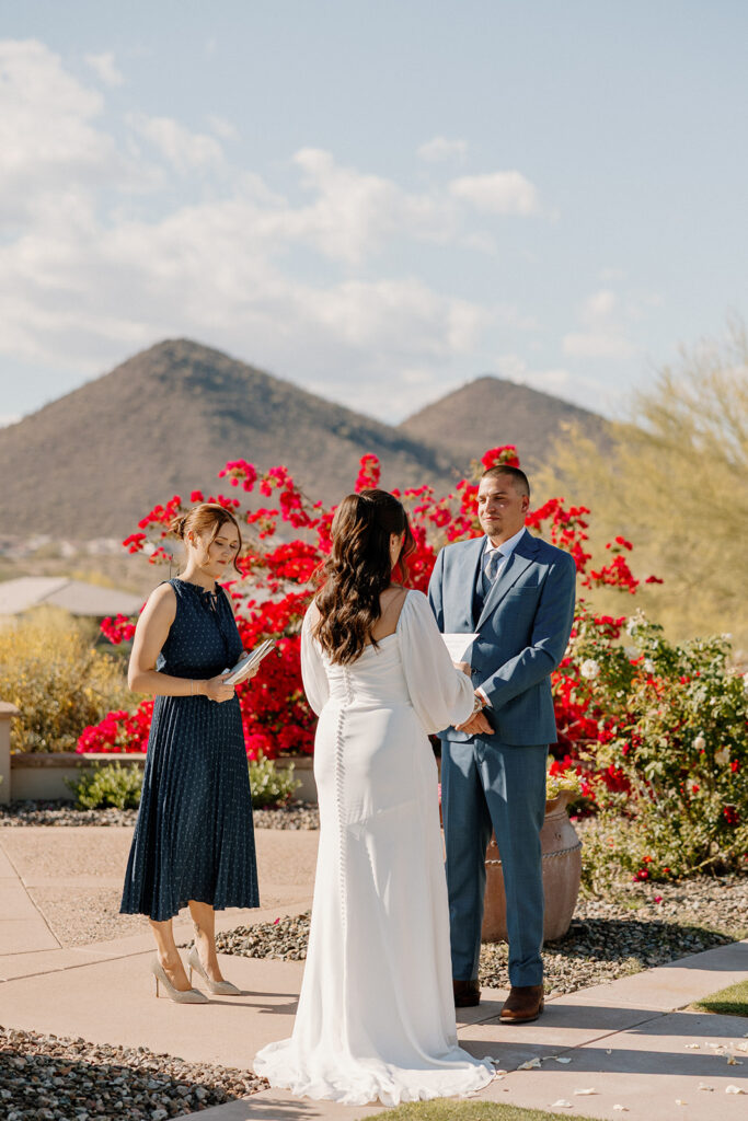 The couple stands in front of vibrant desert blooms and mountain peaks, exchanging vows under the Arizona sun at Blackstone Country Club.