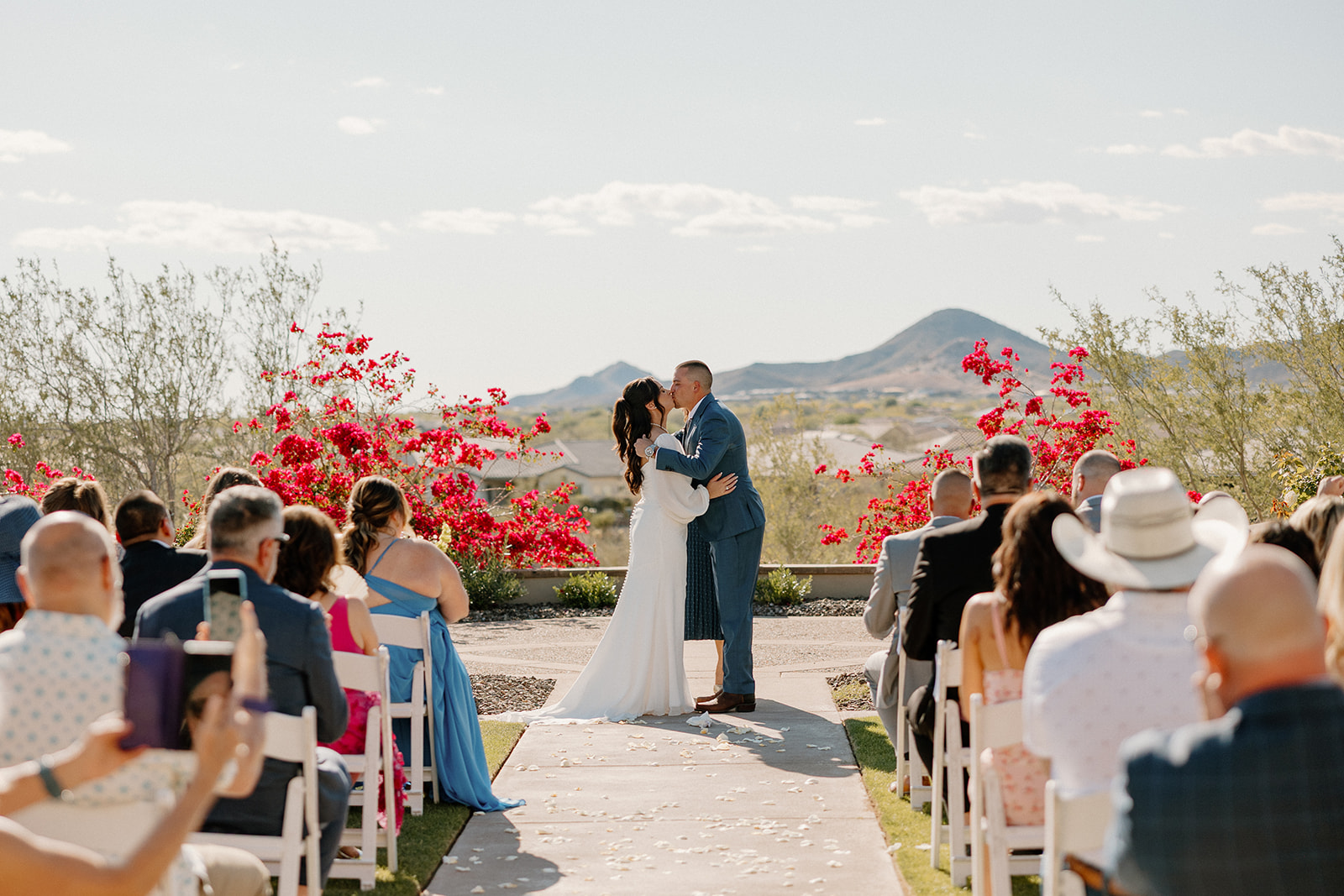 The bride and groom share their first kiss as newlyweds at Blackstone Country Club, framed by red bougainvillea and a stunning desert mountain backdrop.