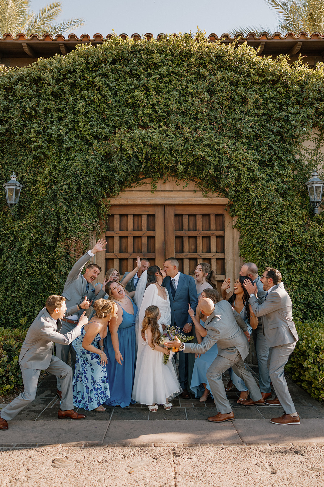 The full wedding party cheers and laughs around the bride and groom in front of the lush ivy wall—total hype squad energy.