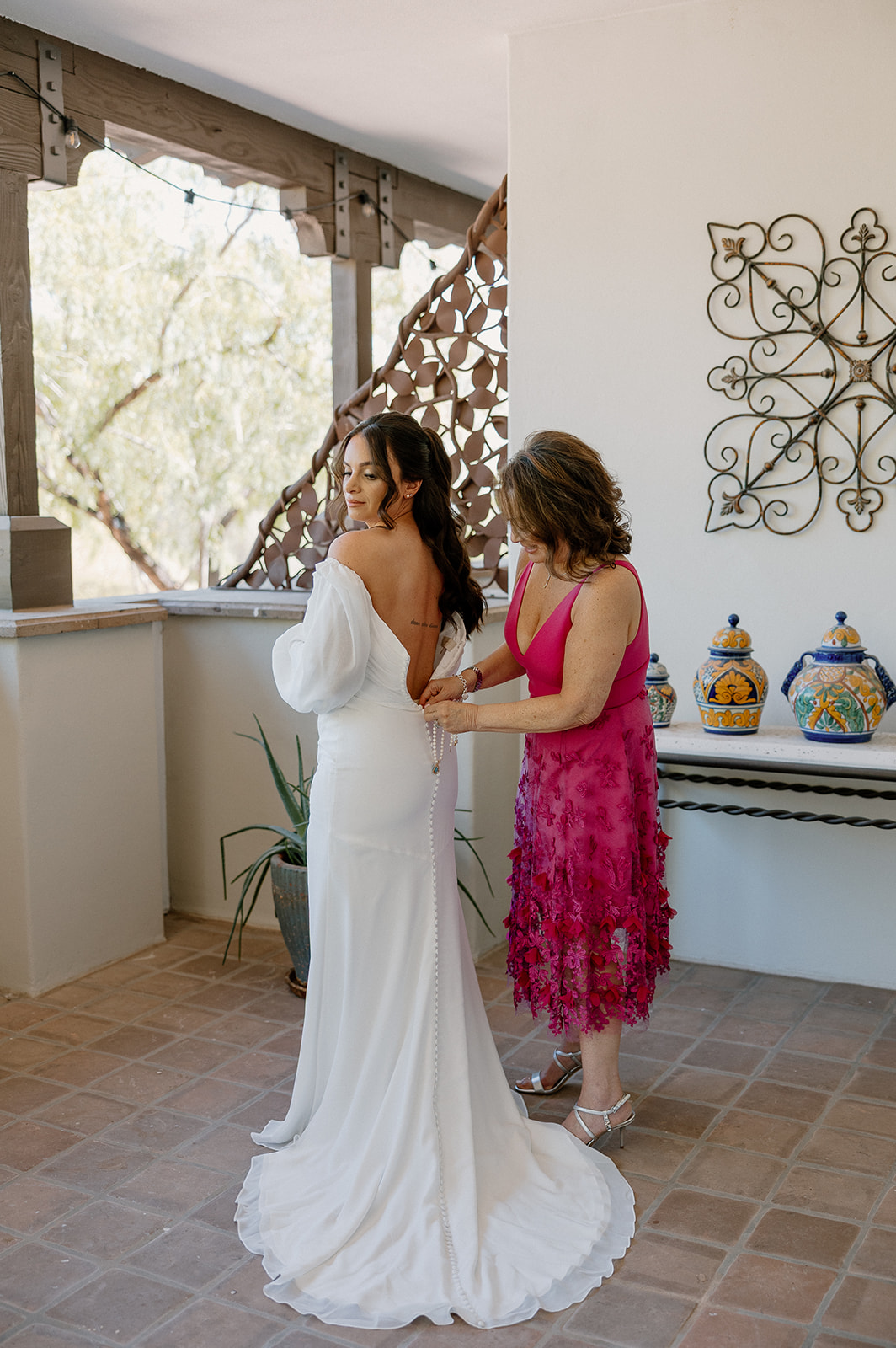 The bride’s mom helps her into her gown, zipping up the back in a bright, airy prep space.