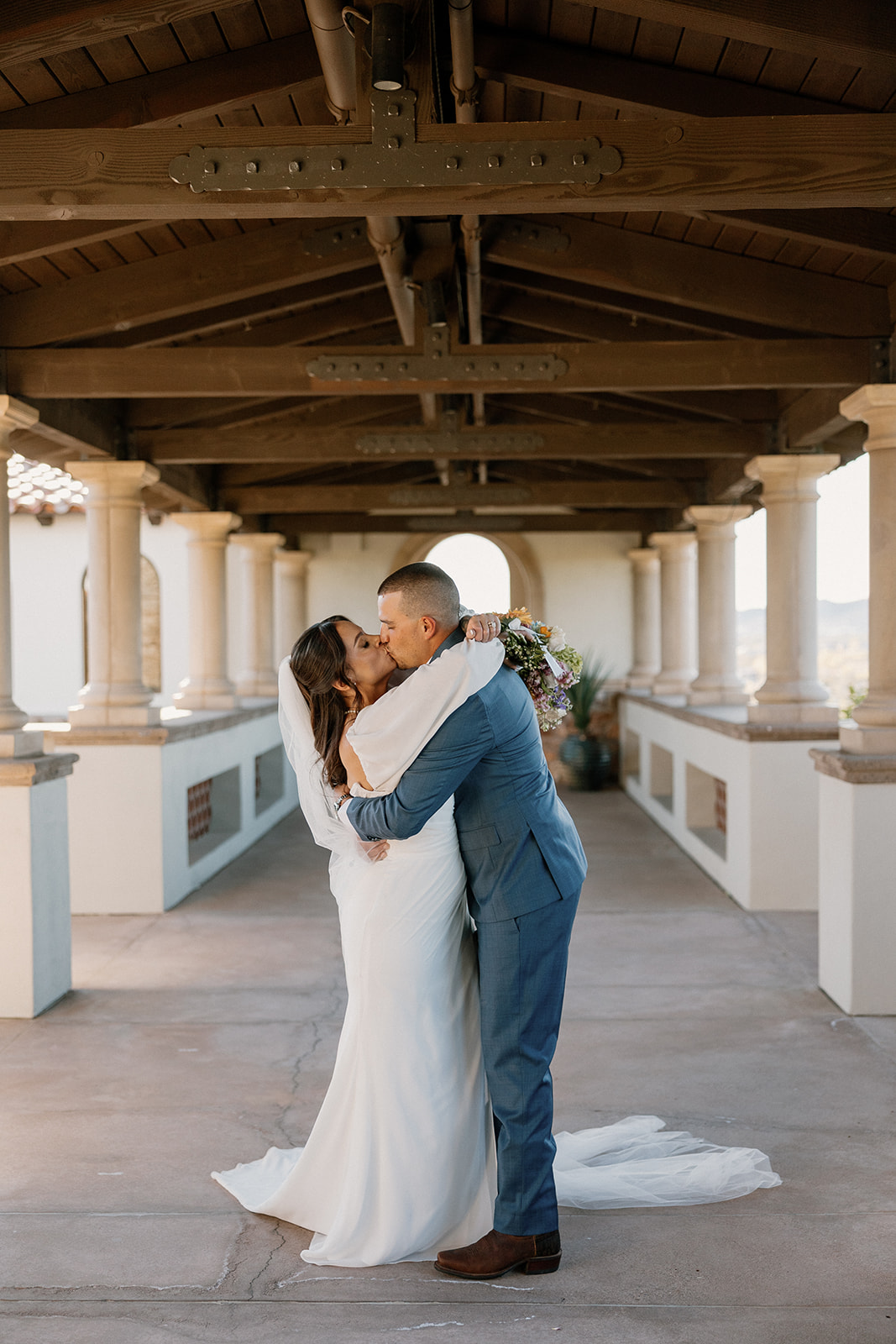 The couple kisses under the grand breezeway at Blackstone Country Club, wrapped in each other beneath the rustic wooden beams.