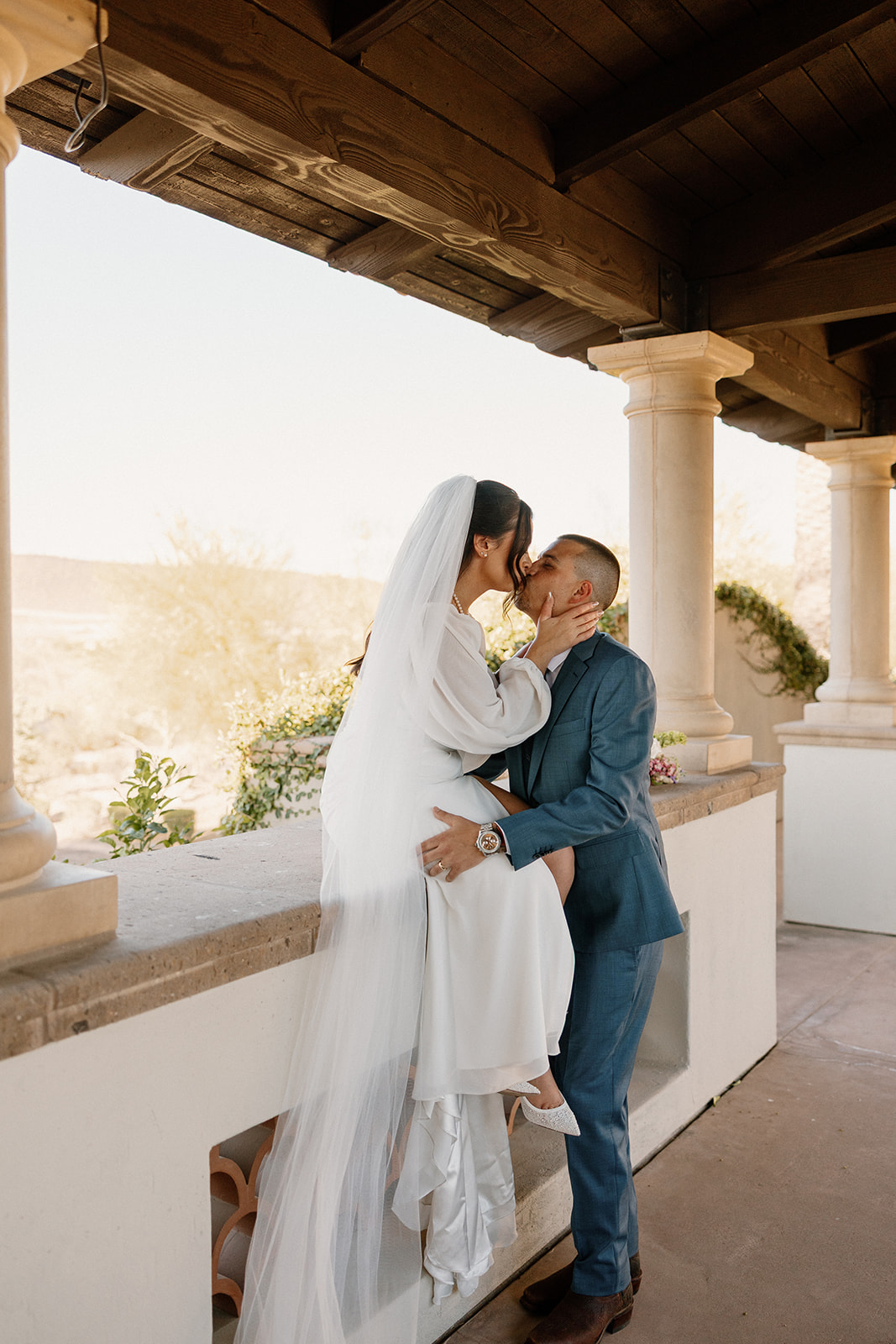 The bride perches on a stucco ledge, pulling her groom in for a kiss at Blackstone Country Club with the desert behind them.