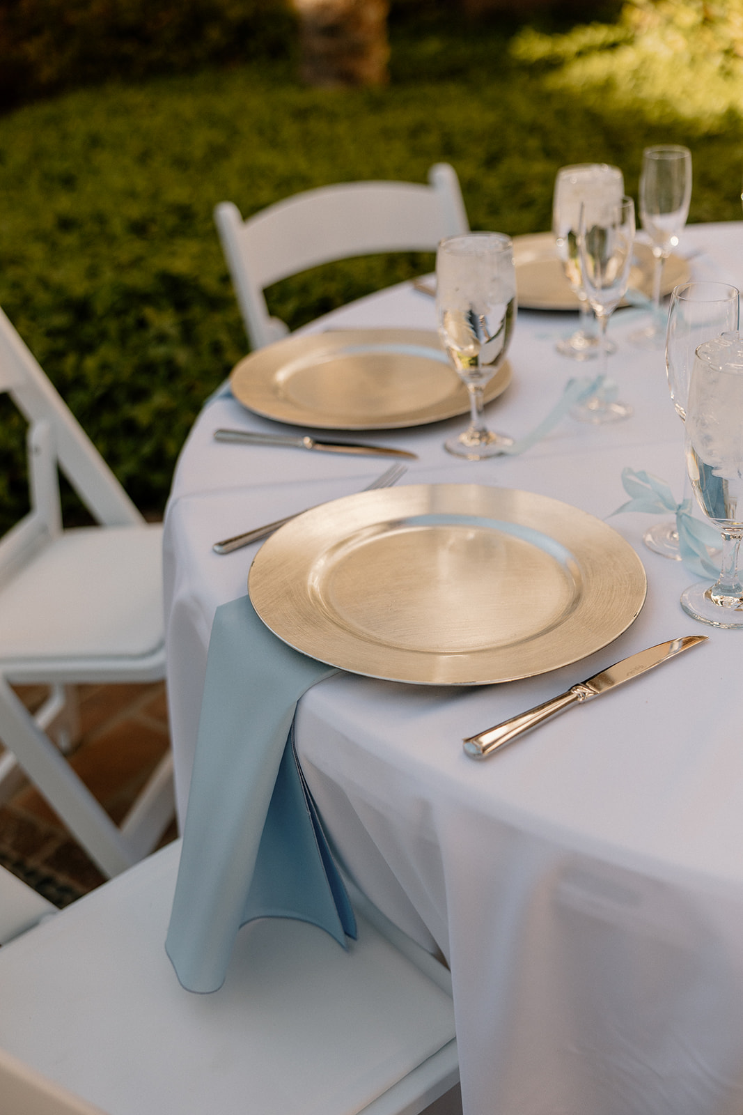 Table details featuring gold chargers, glassware, and soft blue napkins neatly draped over white chairs.