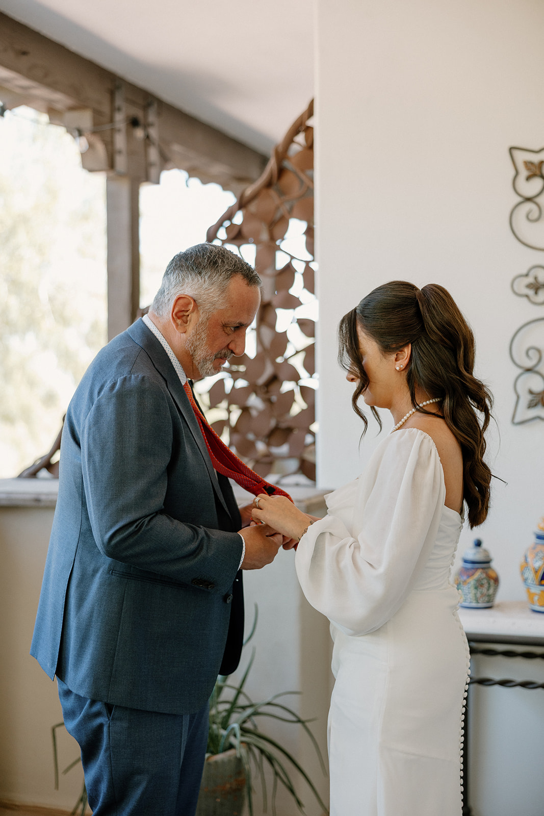 The bride adjusts her dad’s red tie with a sweet smile, surrounded by soft light and sentimental energy.