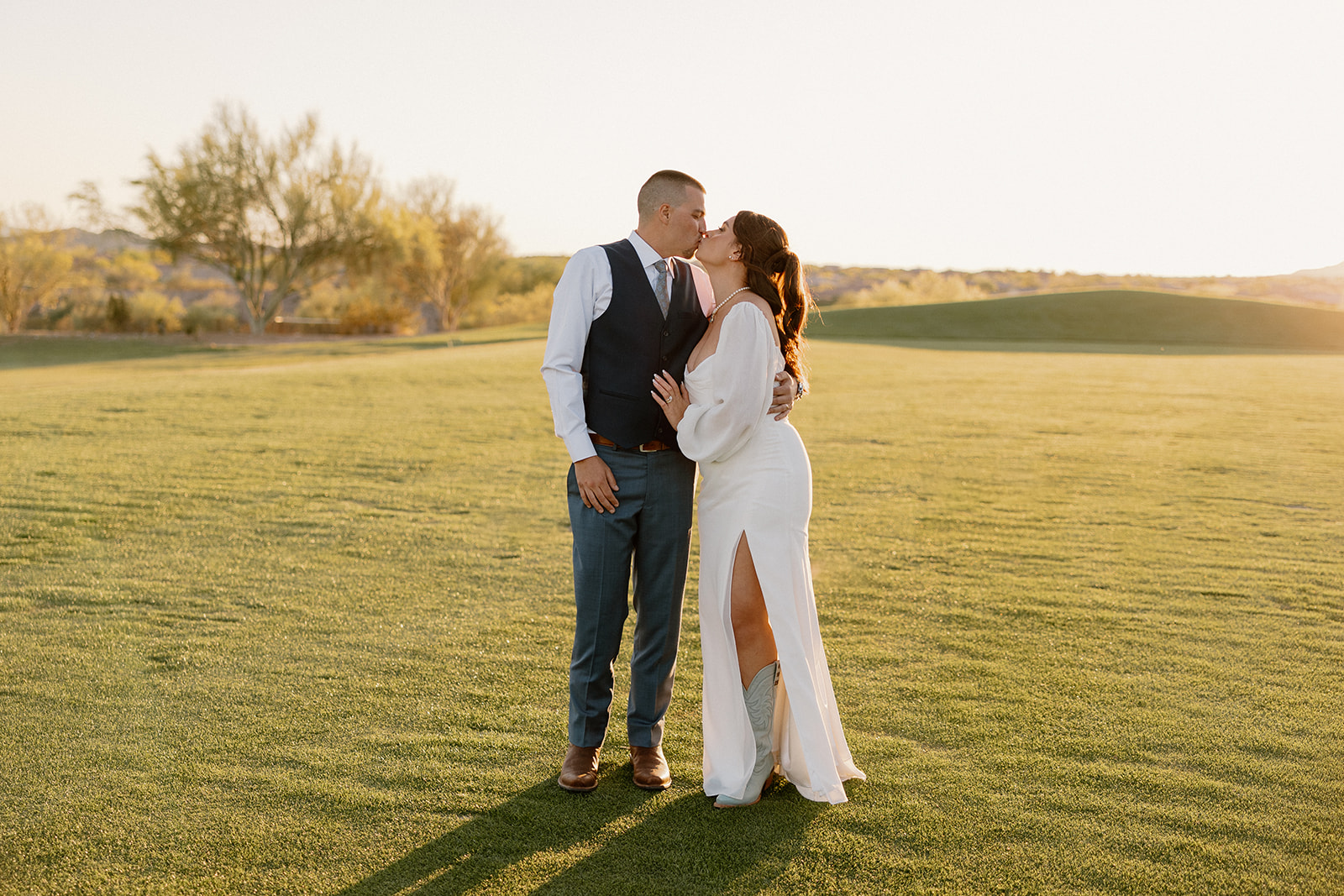 The couple shares a quiet kiss on the lush lawn, backlit by golden light and surrounded by desert calm.