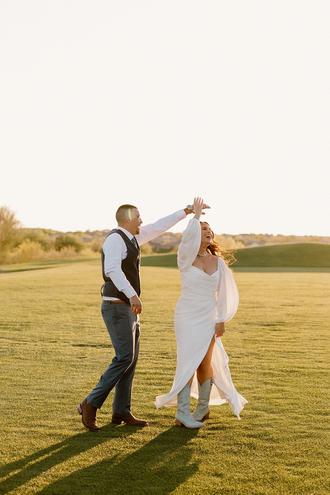 The groom twirls the bride in her long-sleeved gown on the sun-drenched fairway, her boots peeking out with every spin.