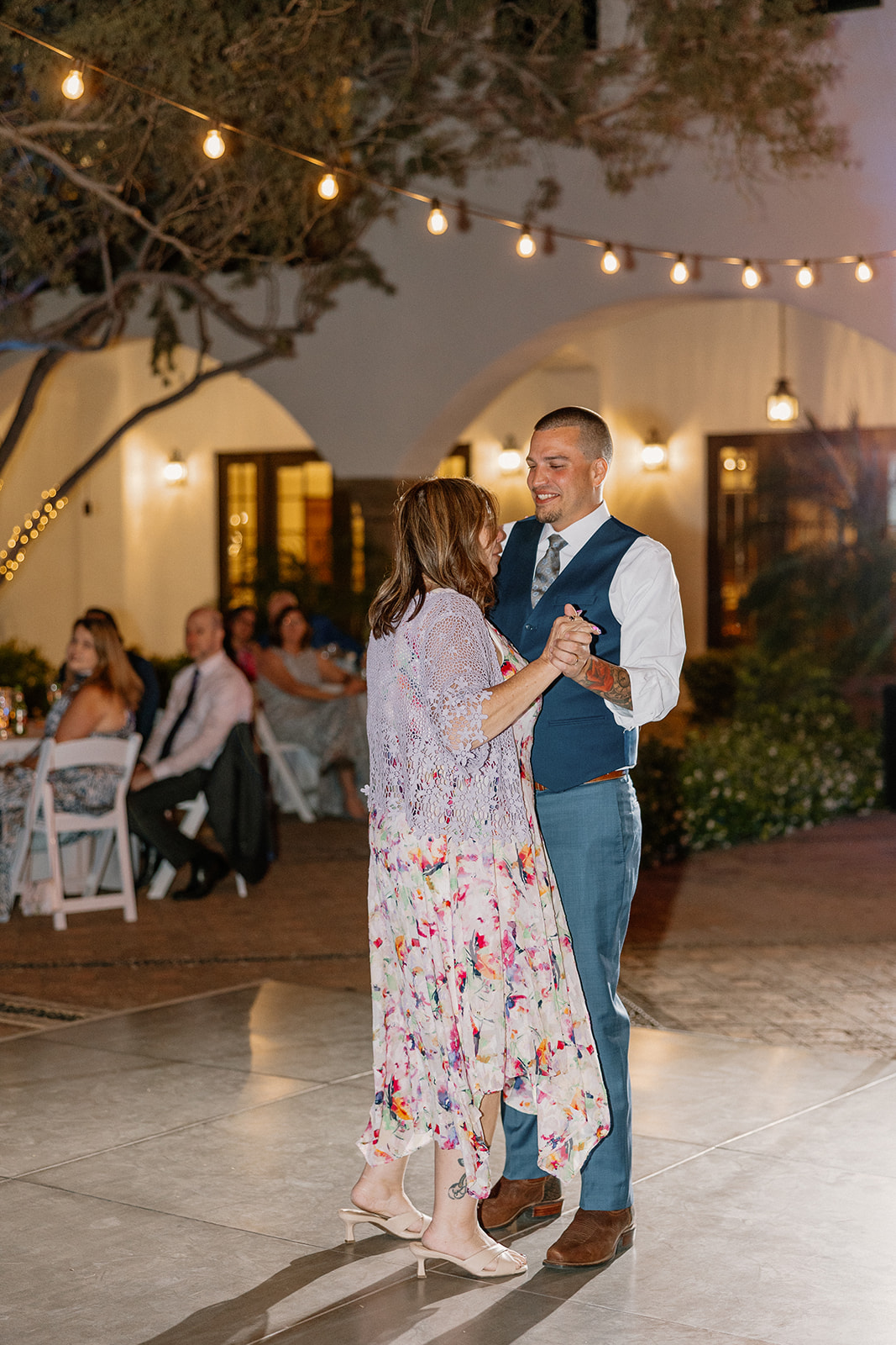 The groom twirls his mother on the outdoor dance floor, warm evening light spilling over the courtyard patio.
