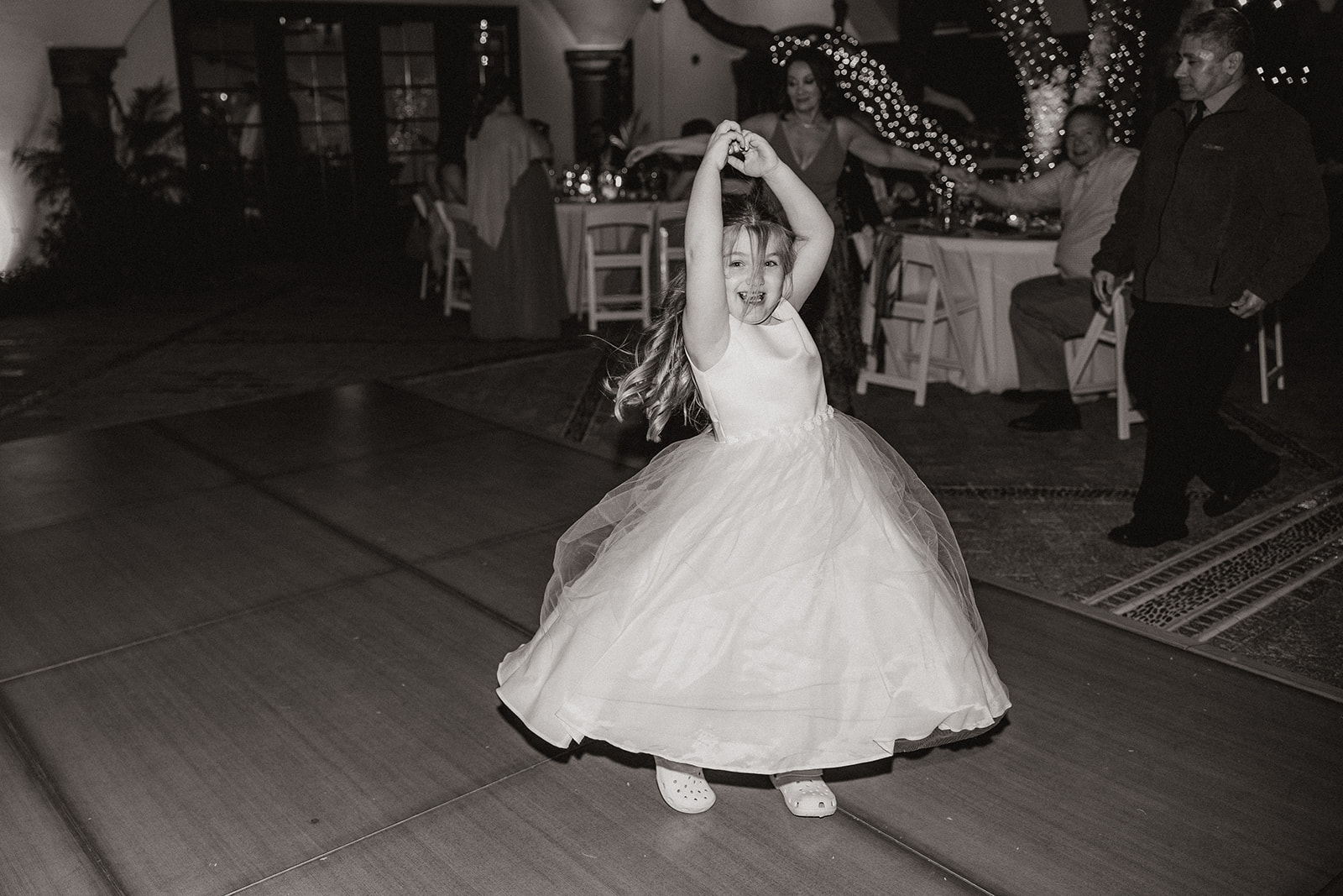 A flower girl in a poofy tulle dress twirls with carefree joy on the dance floor, caught in a candid black-and-white moment.