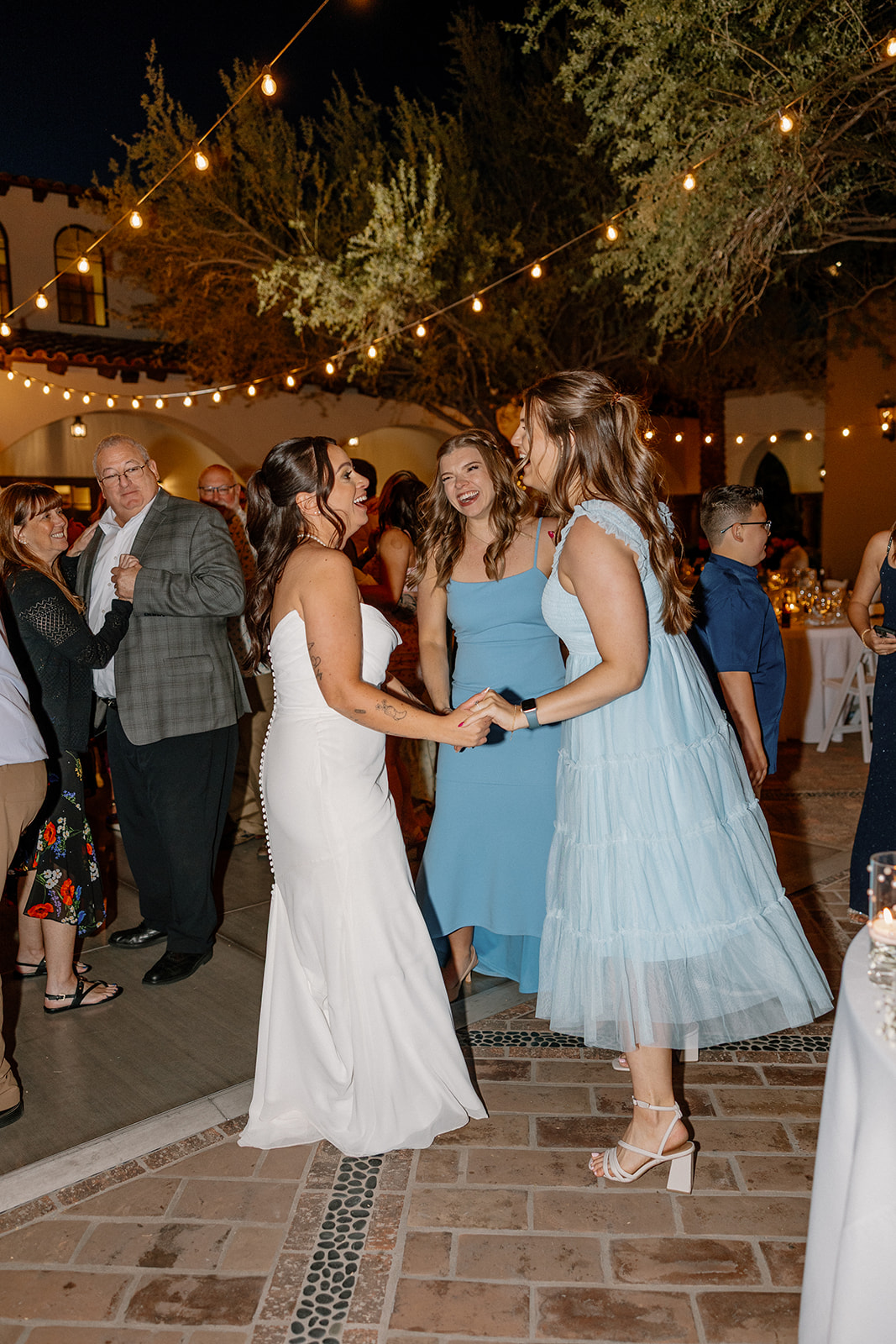 The bride dances barefoot with her bridesmaids in soft blue dresses as laughter and music fill the evening air.
