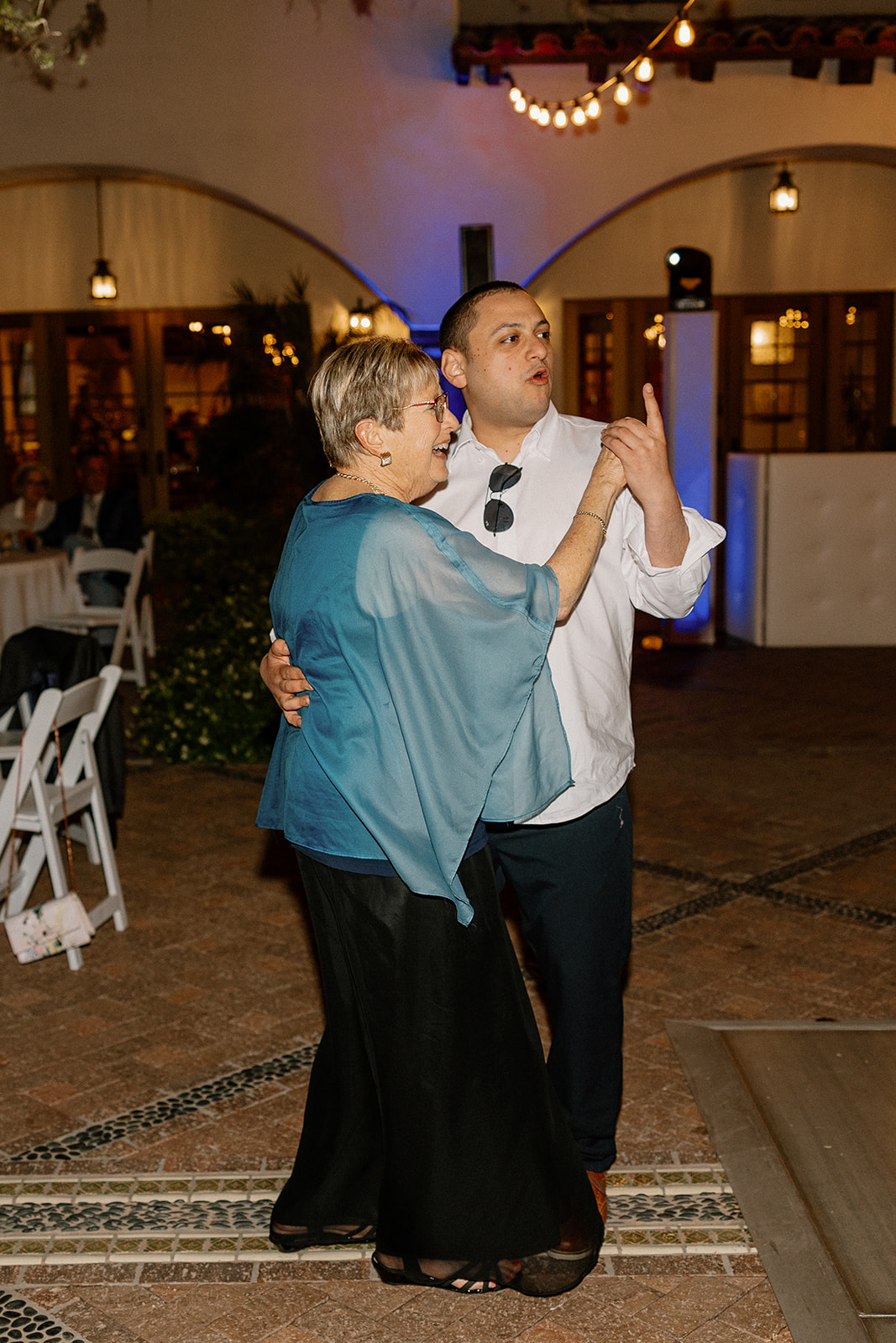 A wedding guest dances with his grandmother on the outdoor patio at Blackstone Country Club, both smiling under string lights.