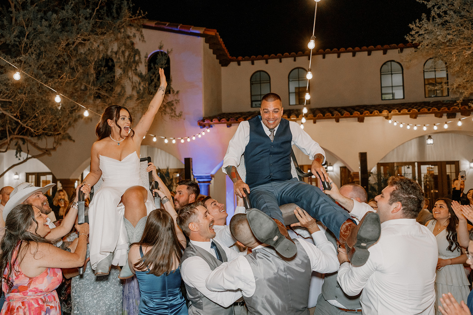 The newlyweds are lifted into the air during the hora dance, surrounded by guests under twinkling string lights at Blackstone Country Club.