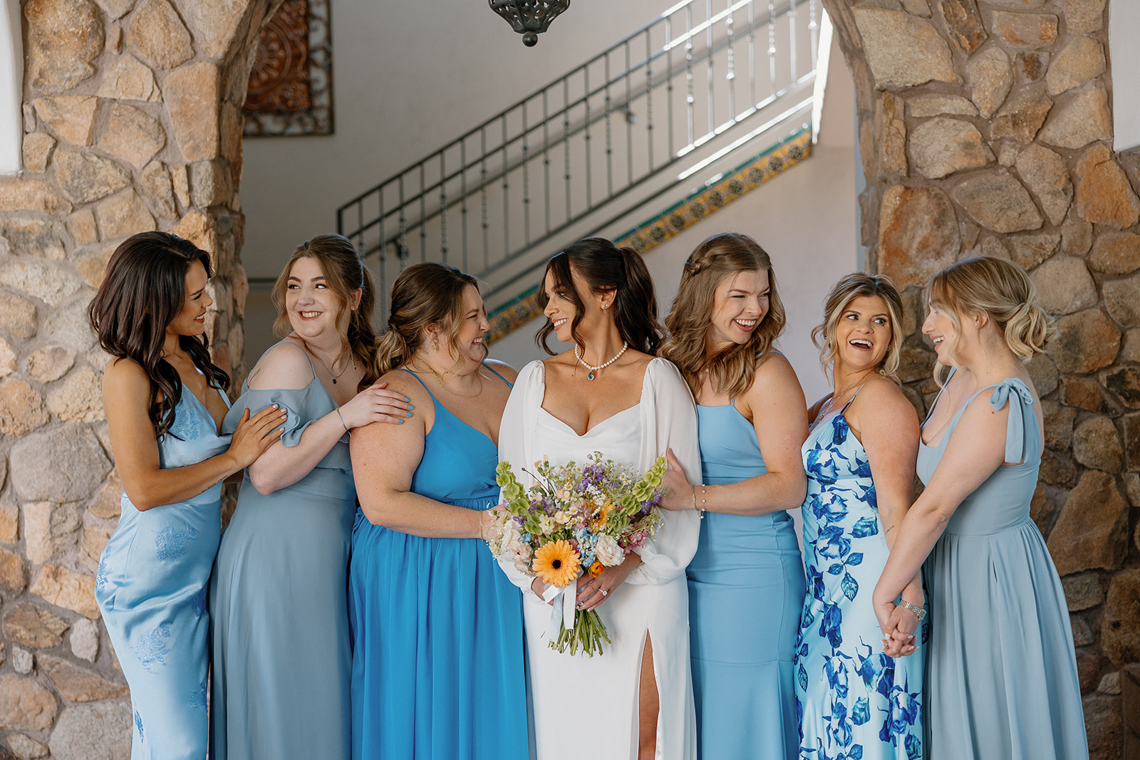 The bride shares a laugh with her bridesmaids in shades of blue, all gathered under a stone archway before the ceremony.