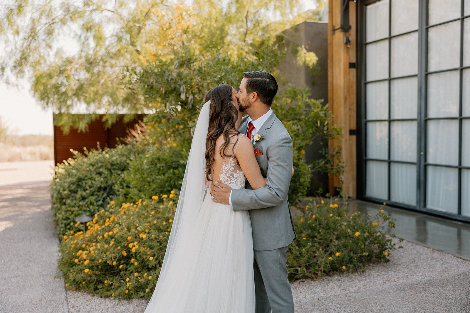 Groom kissing the bride on the temple during their first look outside The Paseo, surrounded by desert blooms.