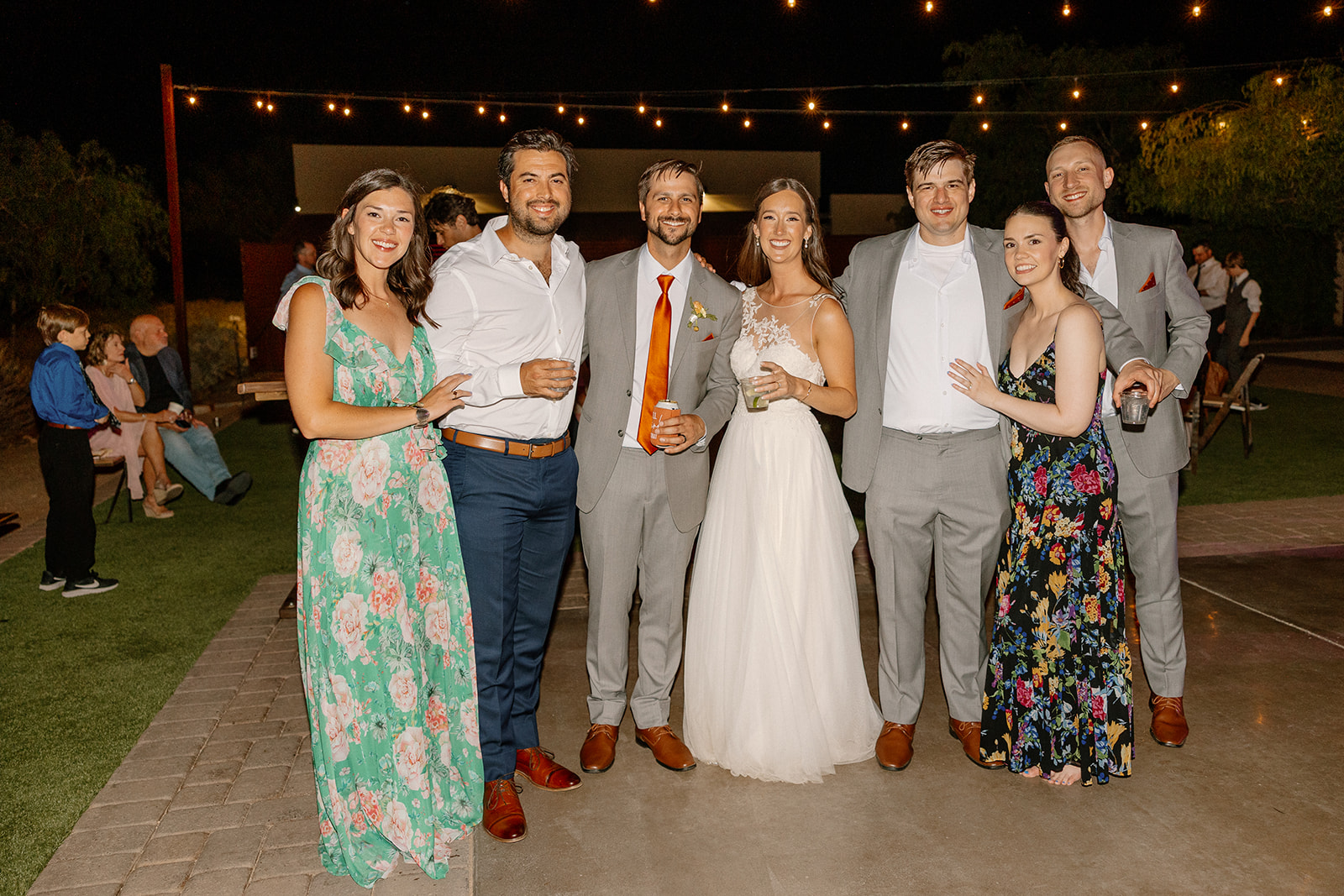 The newlyweds posing with friends under string lights, big smiles and chill vibes all around.