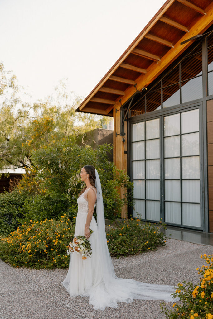 Bride standing in profile with her bouquet outside The Paseo’s reception barn, surrounded by desert blooms.