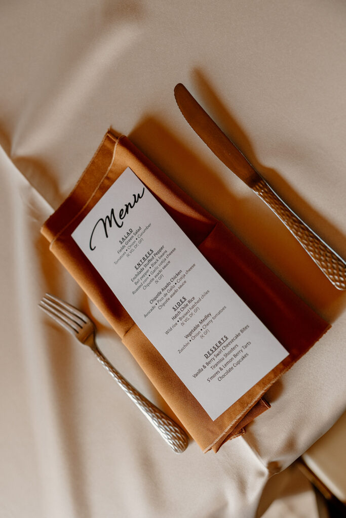 Flat lay of the wedding menu styled with a burnt orange napkin and textured silverware.