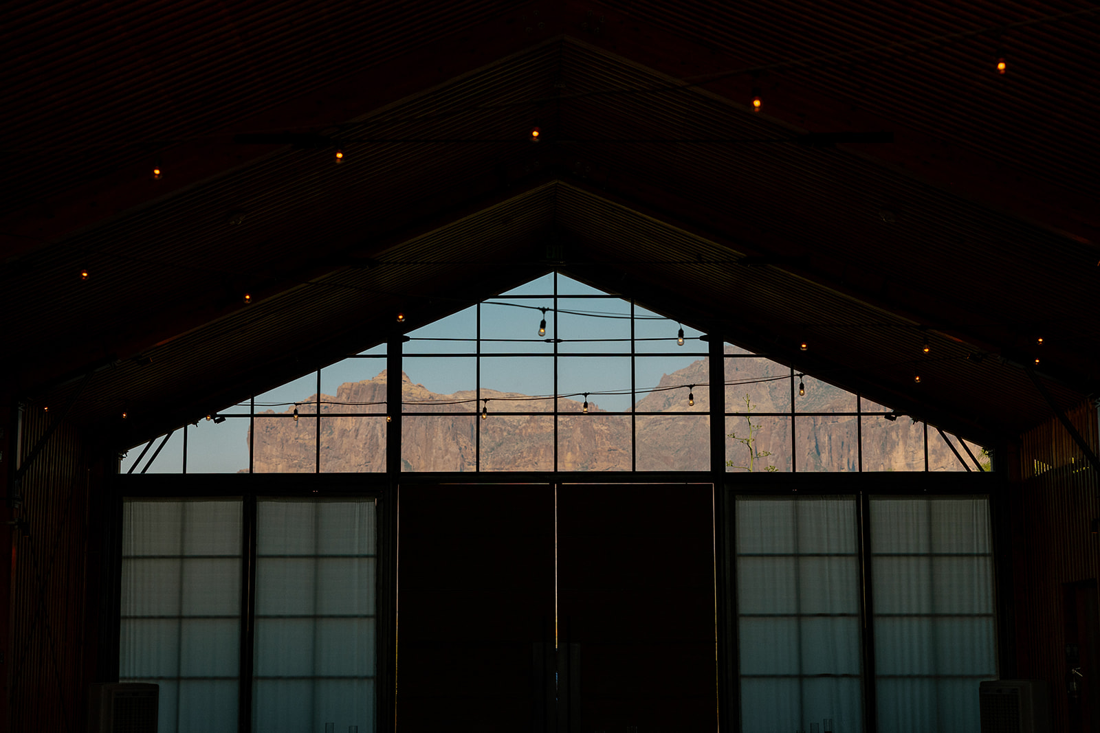 Sunset view of the Superstition Mountains through the glass wall of The Paseo’s reception hall, strung with warm bistro lights.