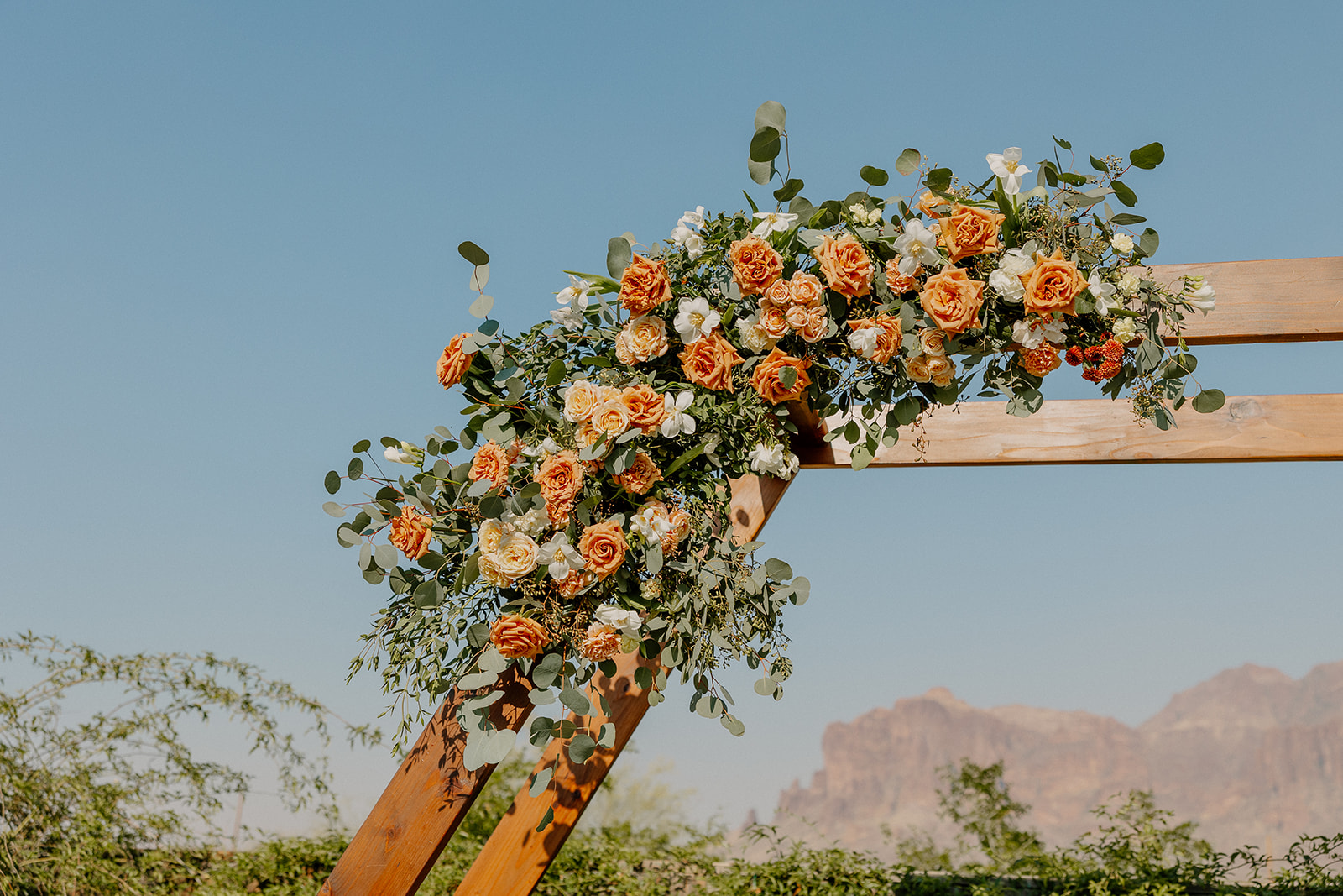 Floral detail on the wooden ceremony arch at The Paseo, with orange and blush roses and the Superstition Mountains behind.