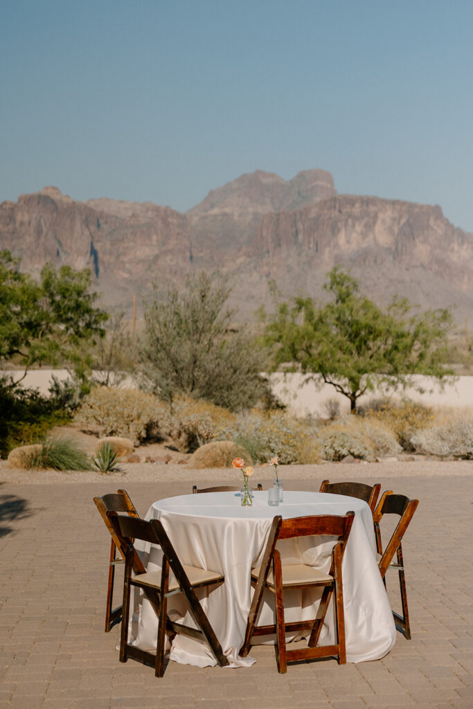 Reception cocktail table set against the Superstition Mountains at The Paseo, with minimalist florals and wood chairs.