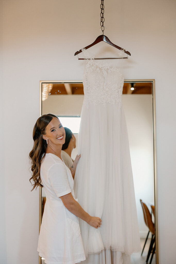 Bride smiling while holding her wedding dress next to a full-length mirror, moments before getting ready.