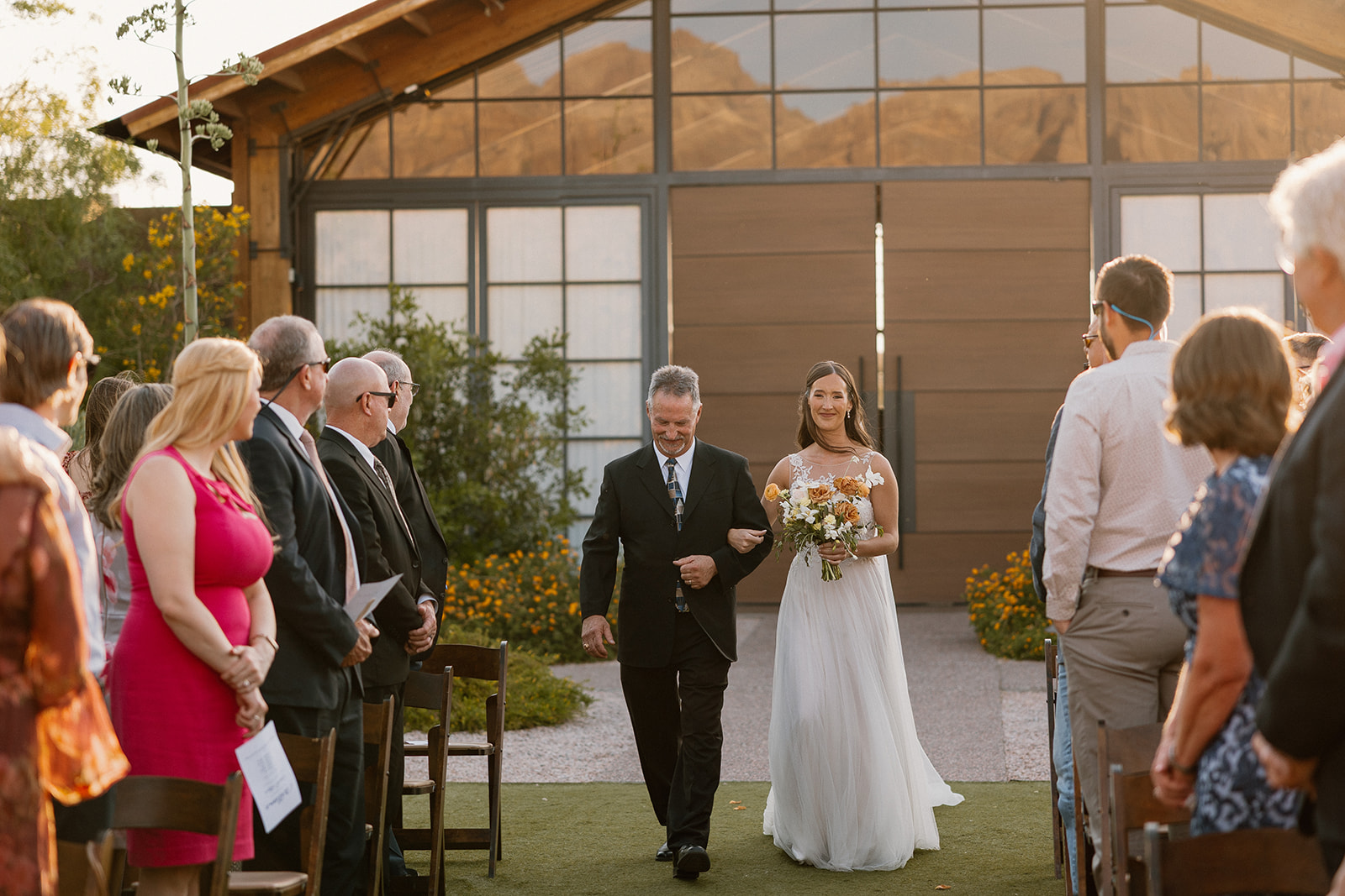 Bride walking down the aisle at The Paseo with her father, guests standing as they approach the ceremony altar.