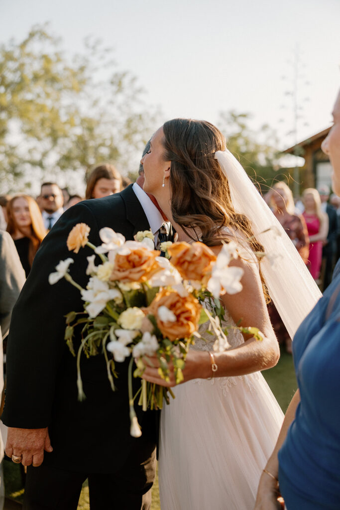 Bride hugging a guest during golden hour, holding a bouquet of peach and white flowers with wild greenery.