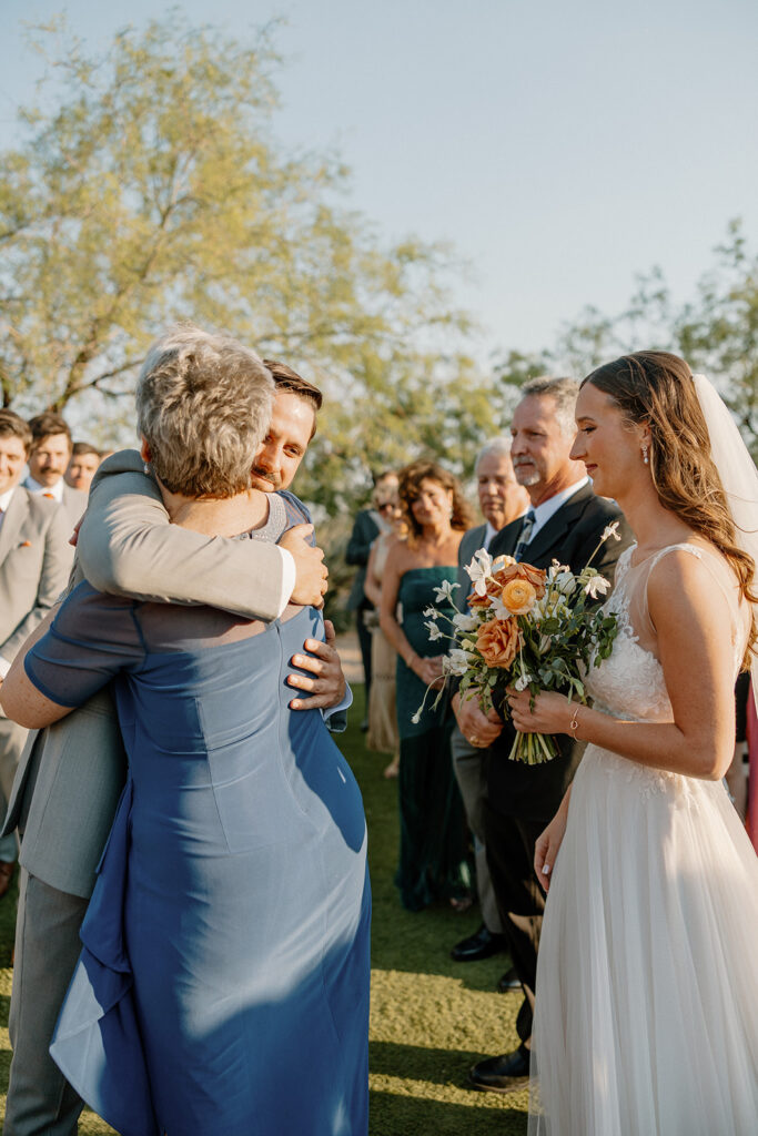 Groom hugging his mom with emotion while the bride looks on, holding her bouquet during the post-ceremony celebration.
