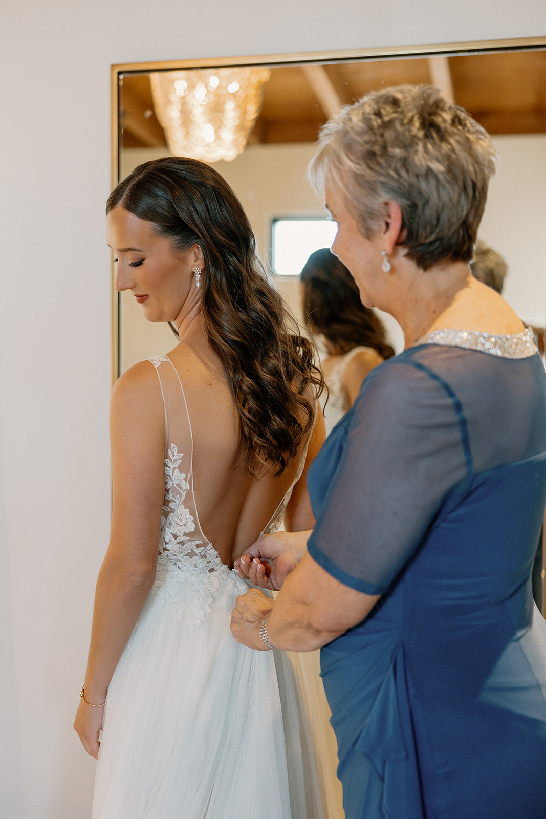 Mother of the bride buttoning the back of her daughter’s gown, soft light and mirror reflections capturing the moment.
