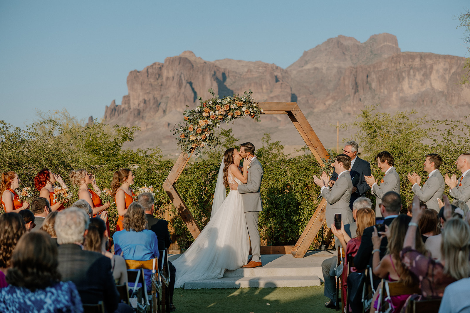 Bride hugging a guest during golden hour, holding a bouquet of peach and white flowers with wild greenery.