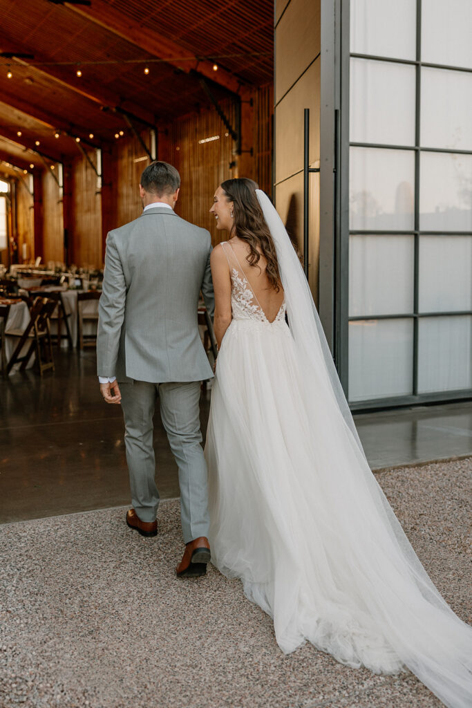 Bride and groom walking hand in hand into The Paseo’s reception barn, soft string lights glowing inside.