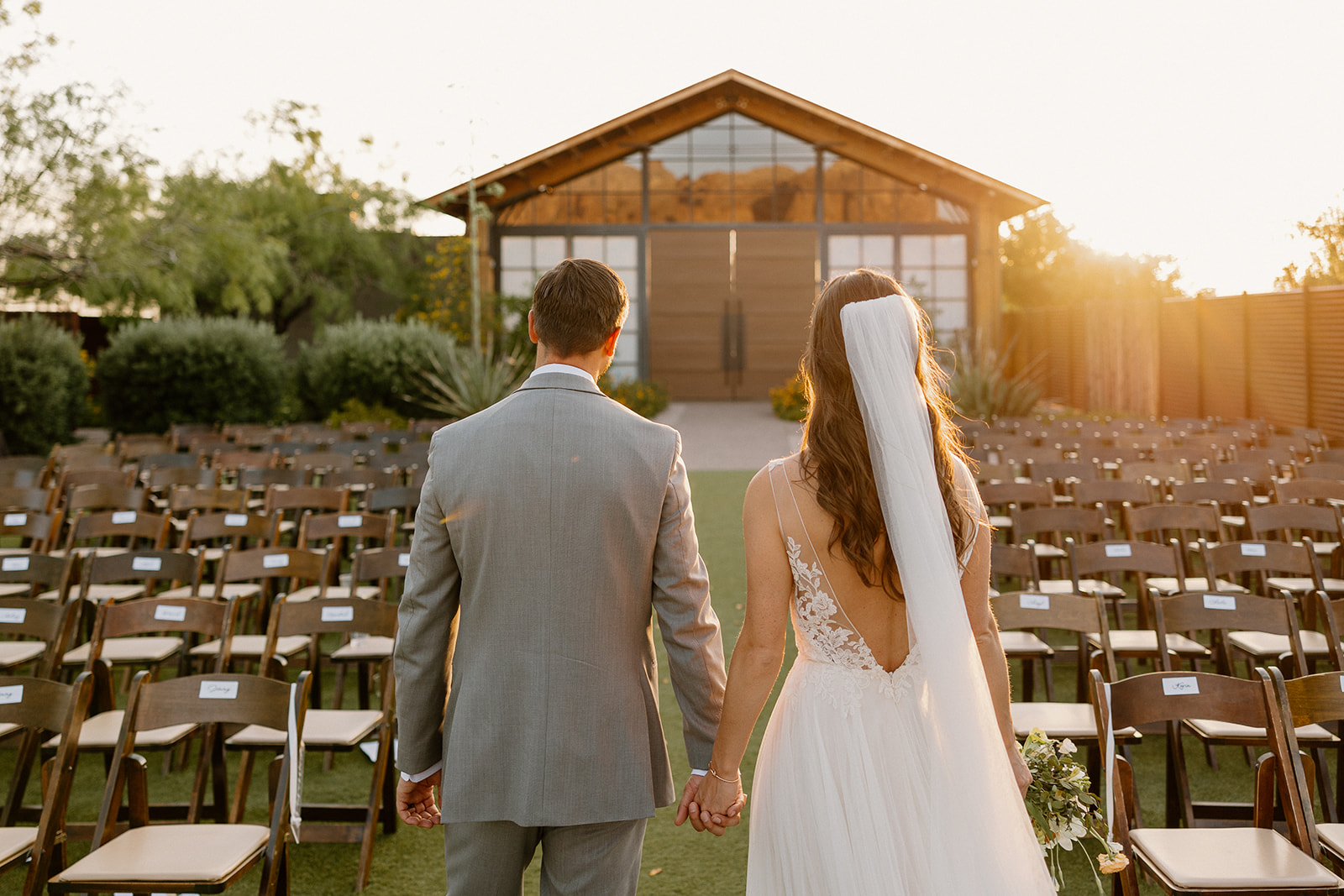 Bride and groom holding hands and walking through the ceremony aisle at golden hour, the reception barn glowing at The Paseo.