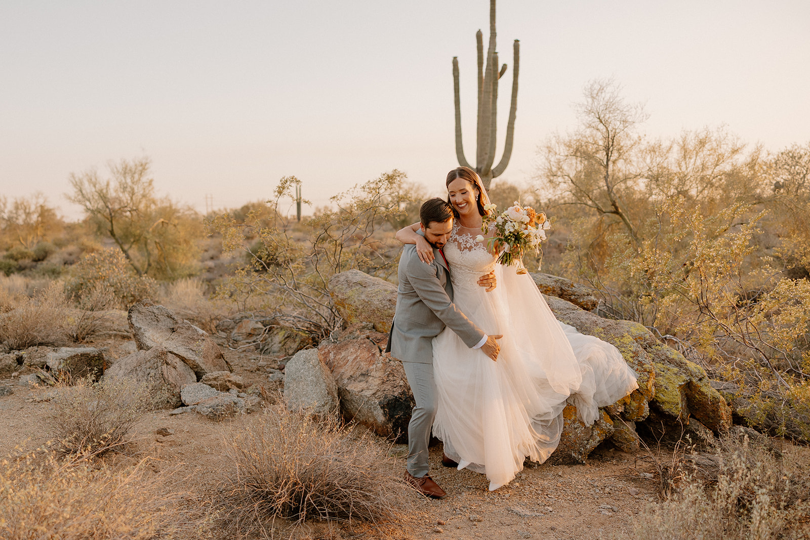 Groom lifting the bride off the rocks in the desert, both laughing with a giant saguaro in the background.