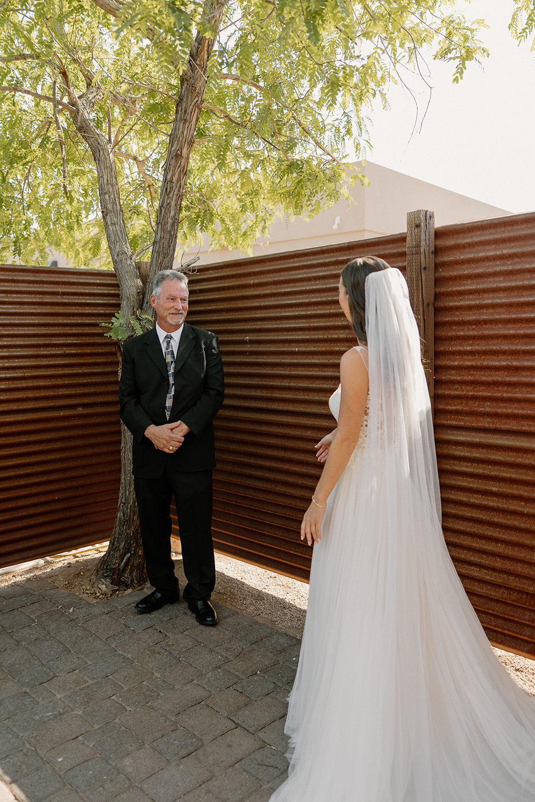 Bride walking toward her father for a first look in a quiet outdoor corner, sunlight filtering through the trees.