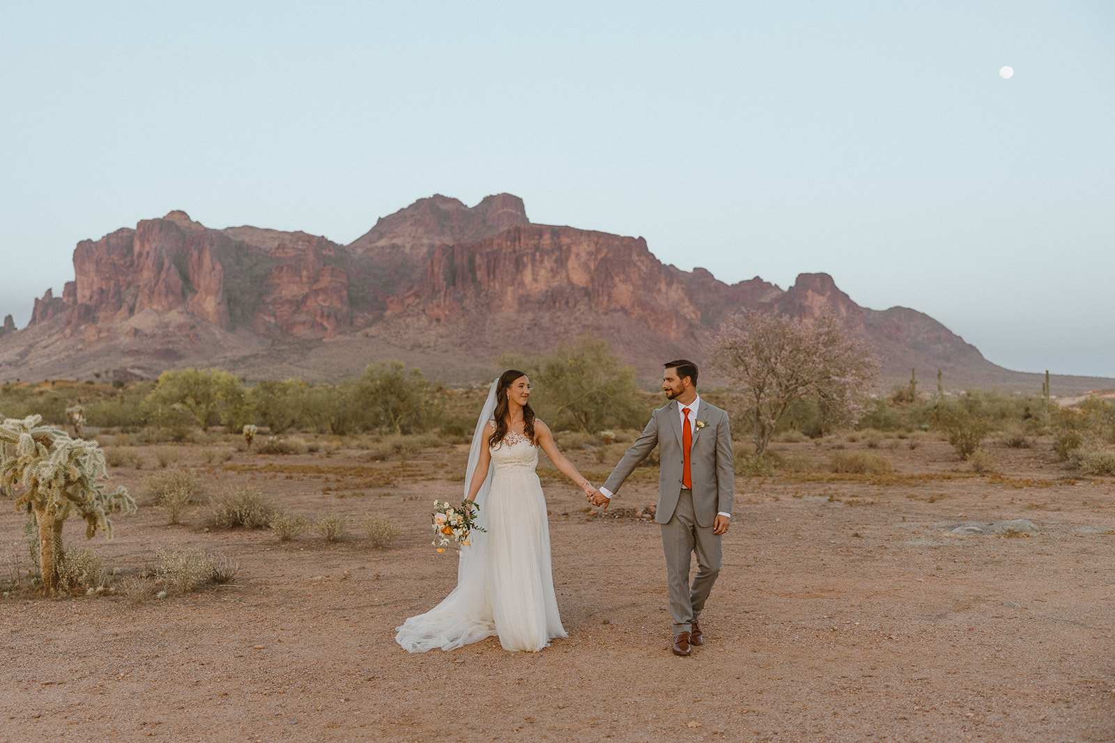 Newlyweds walking hand-in-hand through the desert landscape with the Superstition Mountains in the background at dusk.