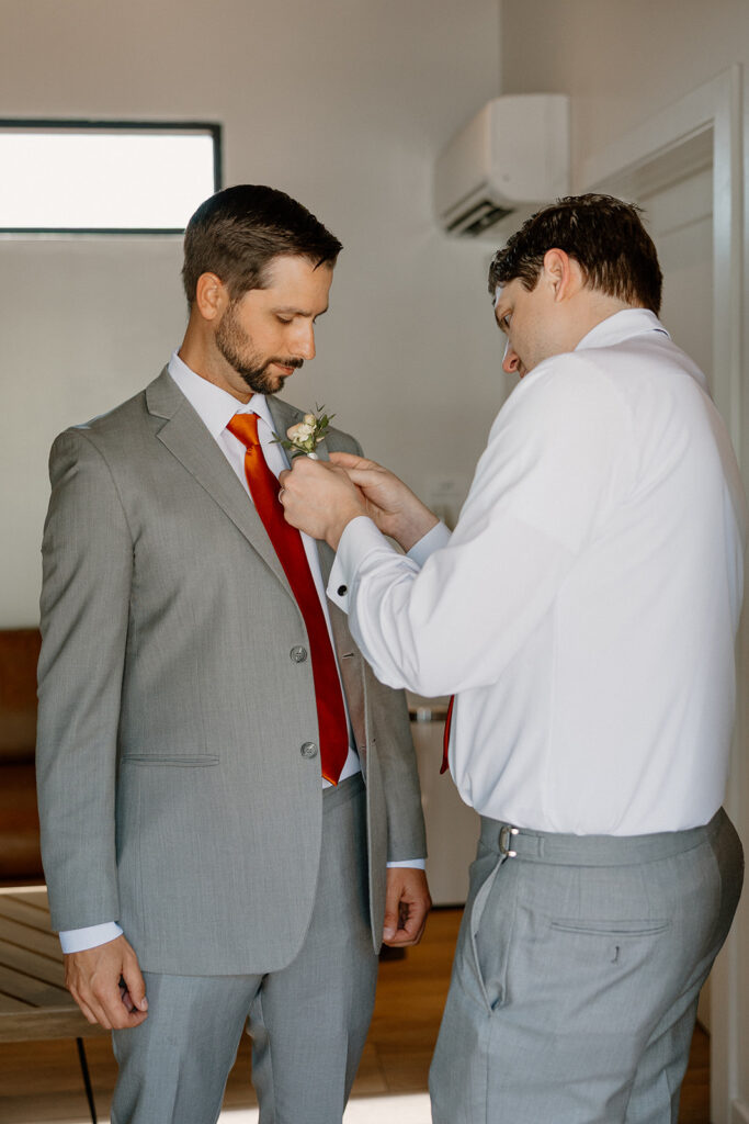 Groomsman helping pin the boutonnière on the groom’s suit before the ceremony.
