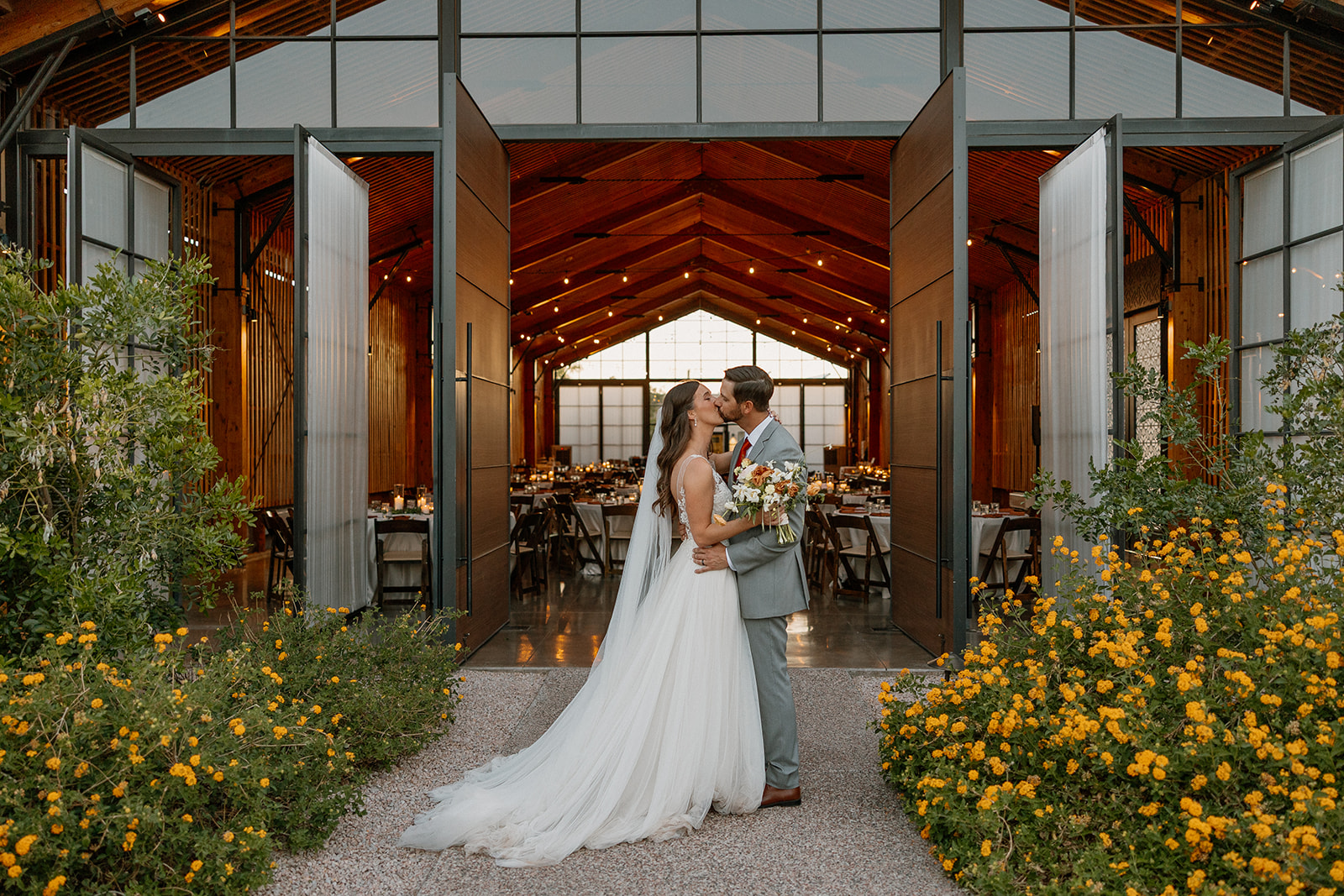 Bride and groom kissing in front of the open reception hall at The Paseo, surrounded by golden wildflowers.