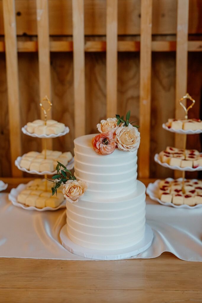 Minimalist white wedding cake with soft florals, surrounded by trays of mini desserts inside The Paseo’s reception hall.