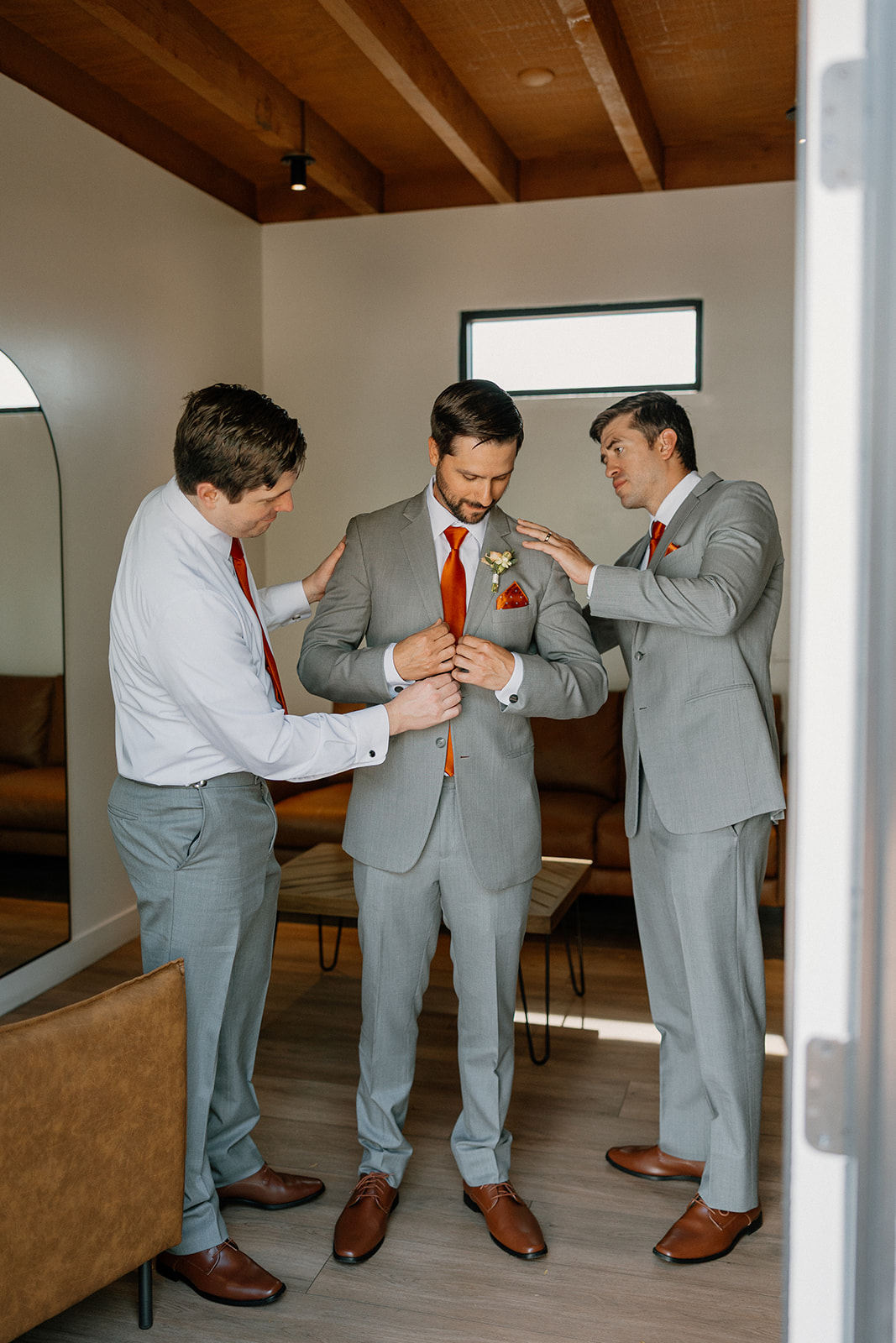 Two groomsmen helping the groom straighten his suit and boutonnière, final touches before heading out.