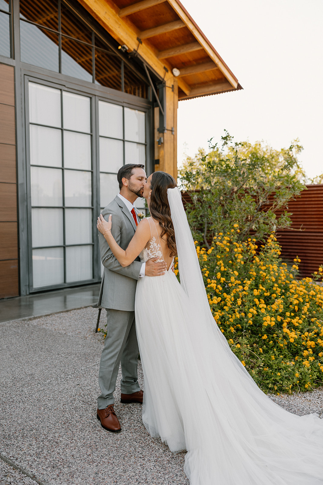 Bride and groom sharing a quiet kiss beside The Paseo’s modern barn, her veil flowing in the breeze.