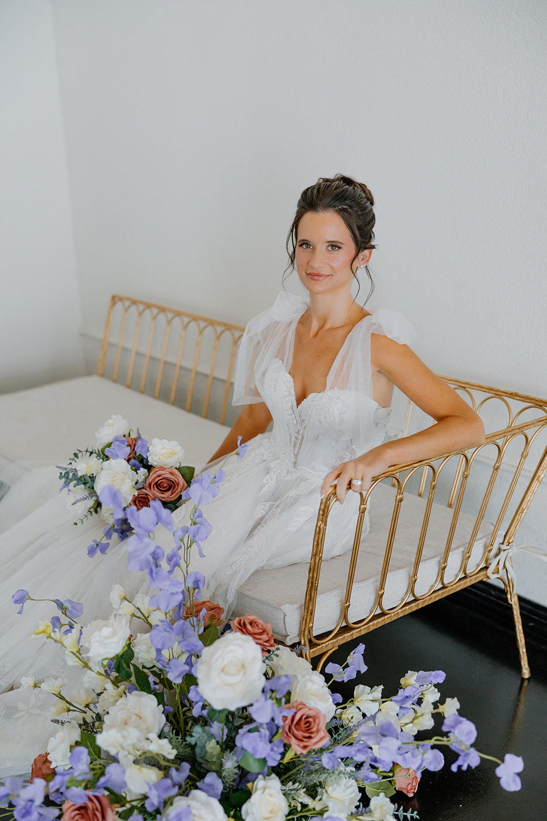 Bride in a romantic wedding gown sitting on a gold bench beside lush pastel florals inside SoHo63.
