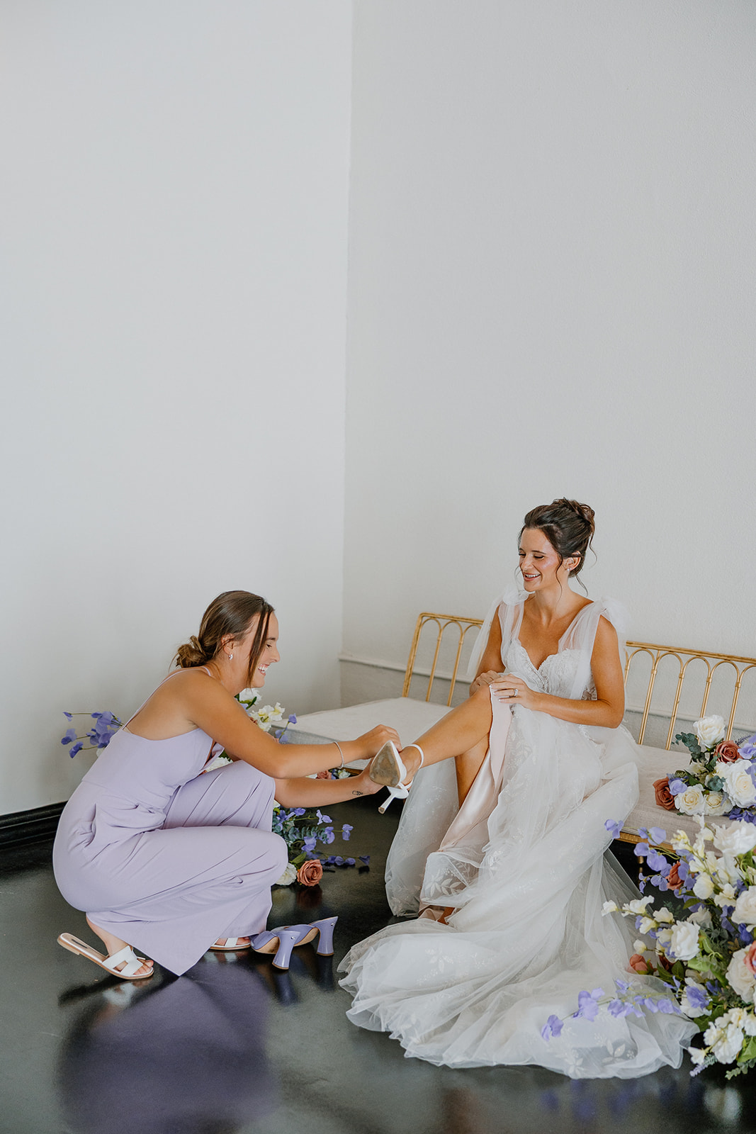 Bridesmaid in lavender jumpsuit helps bride into heels while they laugh in the SoHo63 getting-ready space.