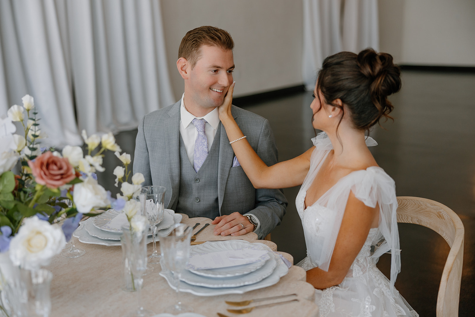 Bride gently touches groom’s cheek as they sit at their styled wedding reception table inside Chandler wedding venue, surrounded by romantic florals.