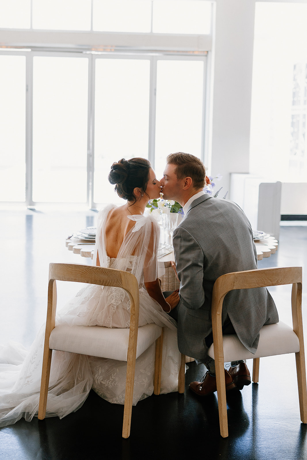 Bride and groom share a kiss while seated at a sweetheart table, framed by soft window light and elegant reception decor inside Chandler wedding venue.