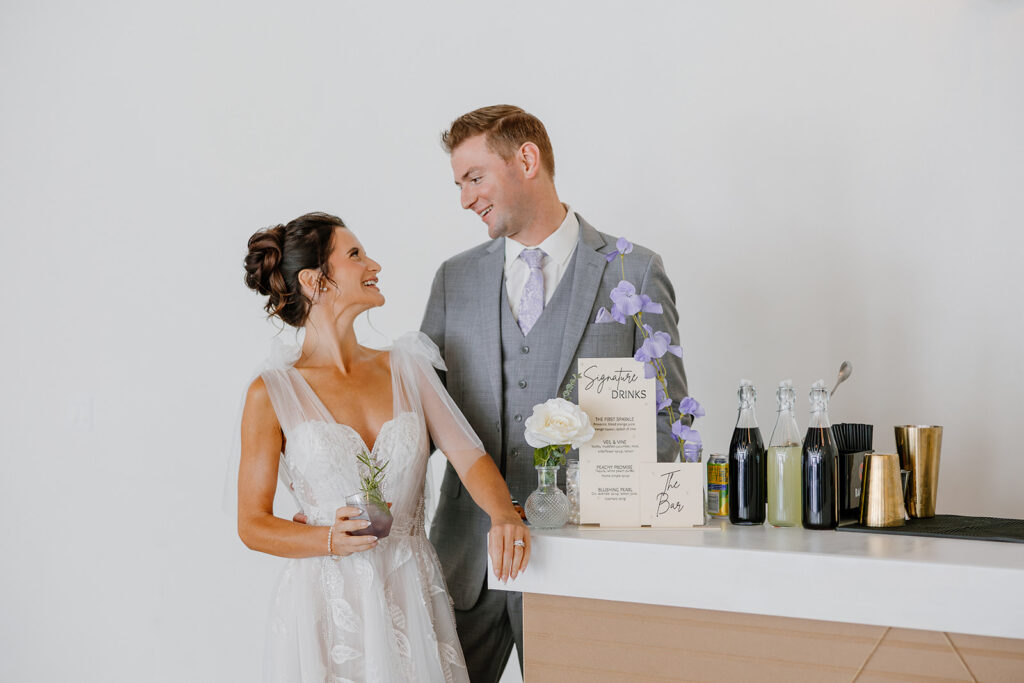 Bride and groom smiling at each other while holding drinks at their custom bar setup inside SoHo63.