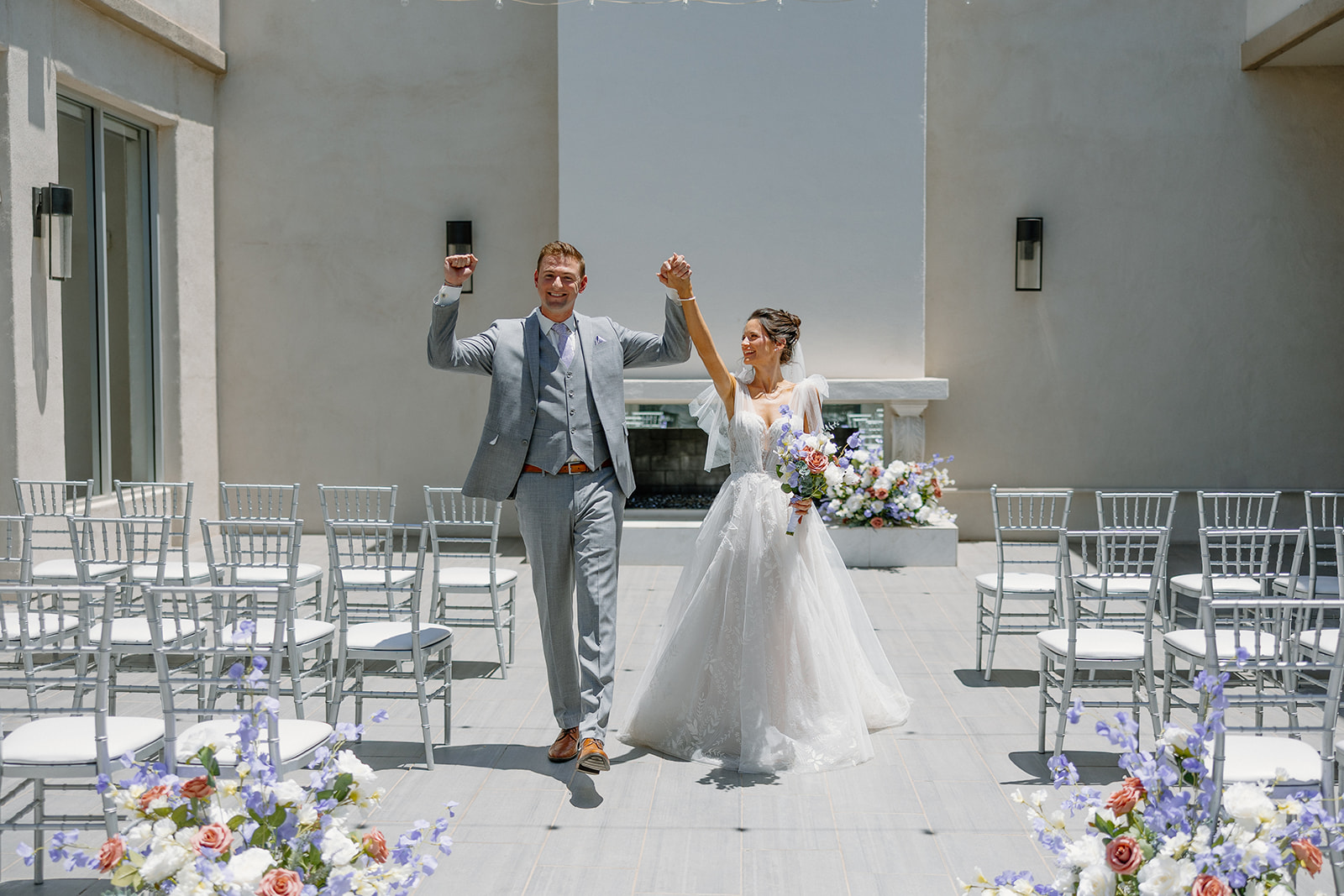 Newlyweds raise their hands in celebration after their Chandler wedding venue ceremony, framed by silver chairs and vibrant floral arrangements.