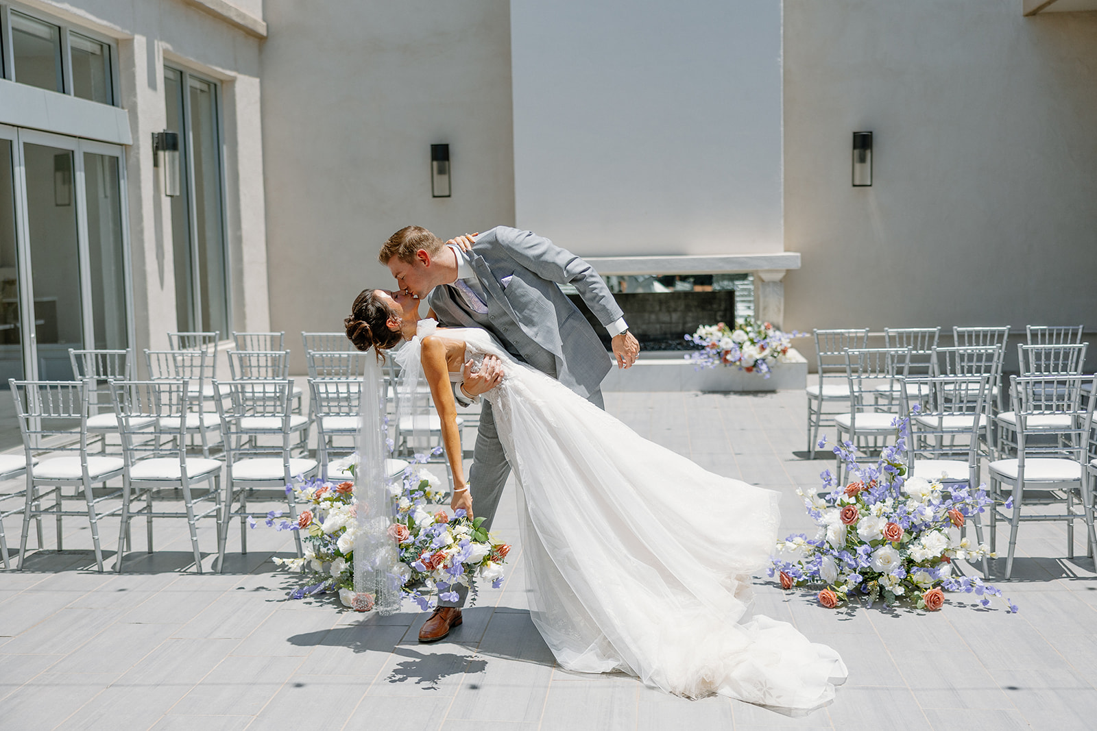 Bride and groom share a romantic dip kiss during their outdoor SoHo63 ceremony with modern fireplace and pastel floral aisle decor.