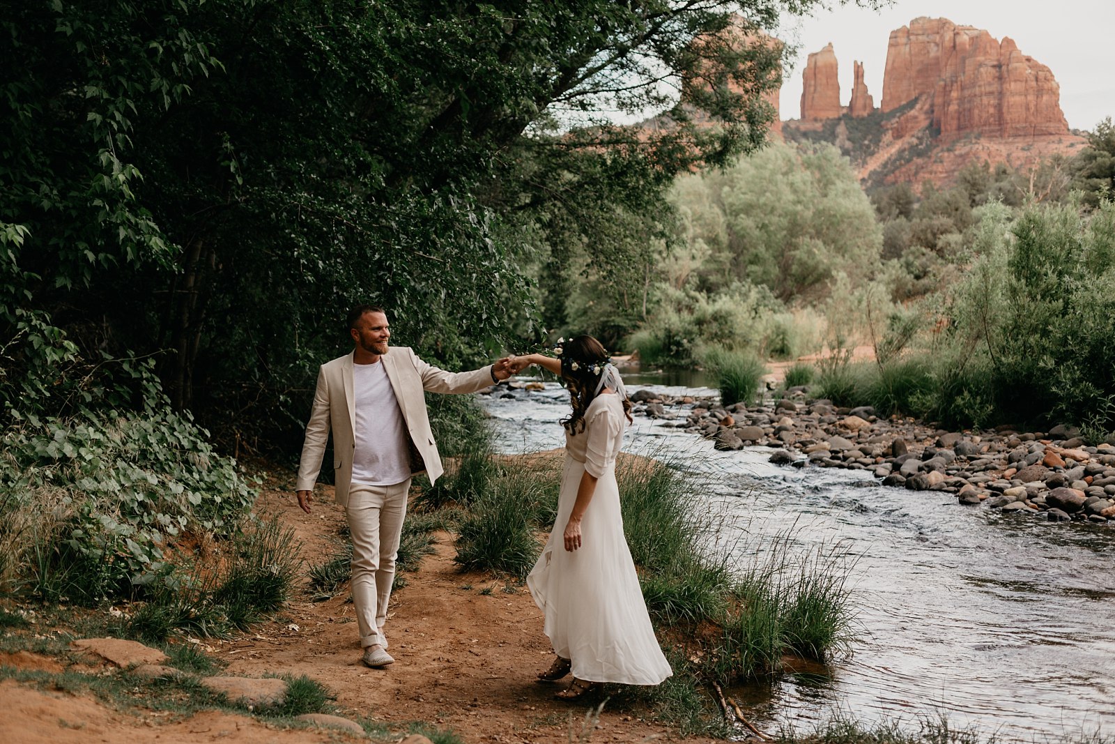 He leads her down a red dirt trail beside the creek, Cathedral Rock glowing in the background during their Sedona elopement.